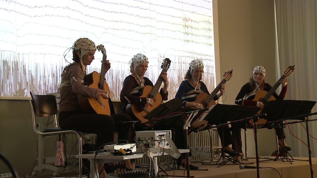 Vier Personen sitzen auf Stühlen und spielen Akustikgitarren. Sie tragen EEG-Hauben, die mit Kabeln verbunden sind. Im Hintergrund ist ein großer Bildschirm mit EEG-Wellenmustern sichtbar. Vor ihnen stehen Notenständer. Der Raum ist schwach beleuchtet, und es gibt technische Geräte auf einem Tisch.
