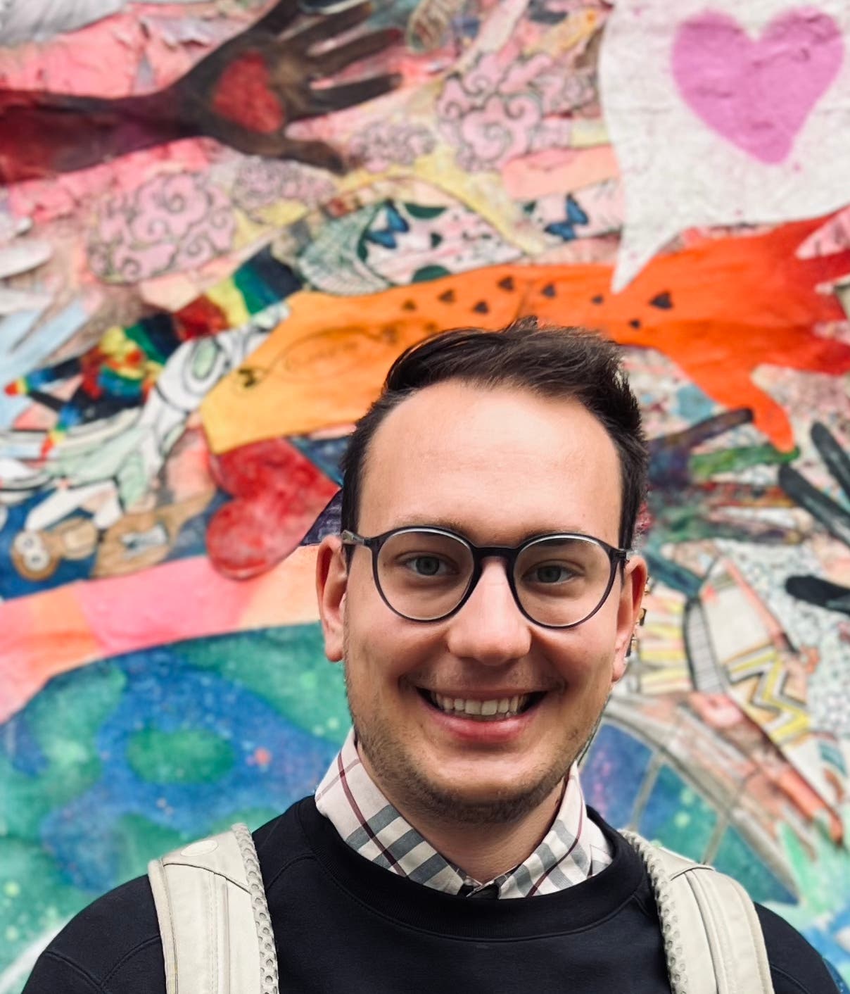 Ein junger Mann mit dunklen Haaren und Brille lacht in die Kamera. Er trägt einen dunklen Pullover, darunter einen karierten Hemdkragen. Im Hintergrund sieht man ein Gemälde von einem bunten Wirrwarr aus Händen und Herzen, darunter ein rosafarbenes Herz in einer Sprechblase, die aus seinem Kopf zu kommen scheint.