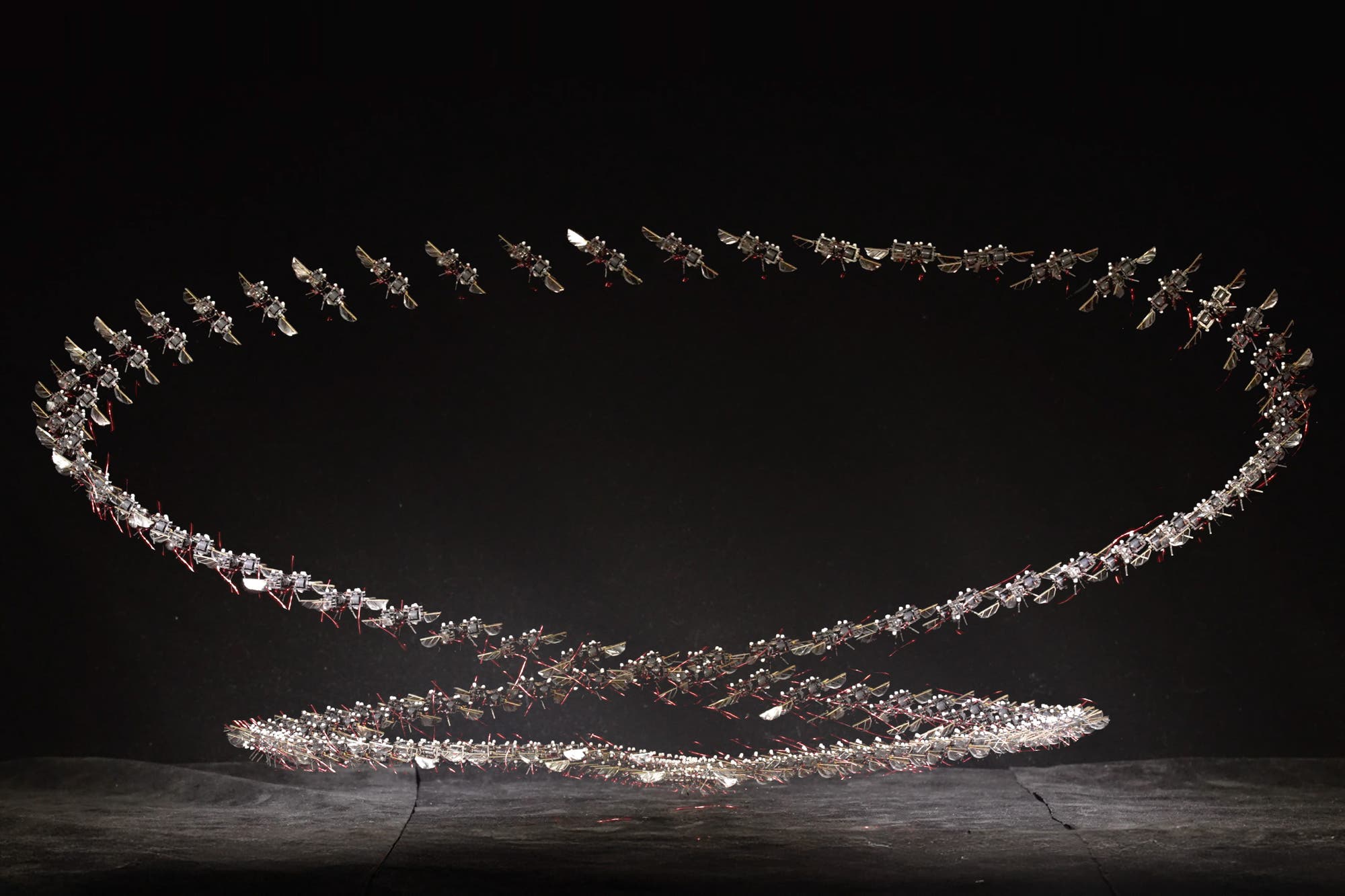 Eine Nahaufnahme eines kleinen, mechanischen Geräts mit Flügeln, das auf einer menschlichen Hand ruht. Das Gerät hat eine komplexe Struktur mit sichtbaren Drähten und mechanischen Komponenten, die an Insektenflügel erinnern. Die Hand dient als Maßstab, um die geringe Größe des Geräts zu verdeutlichen.