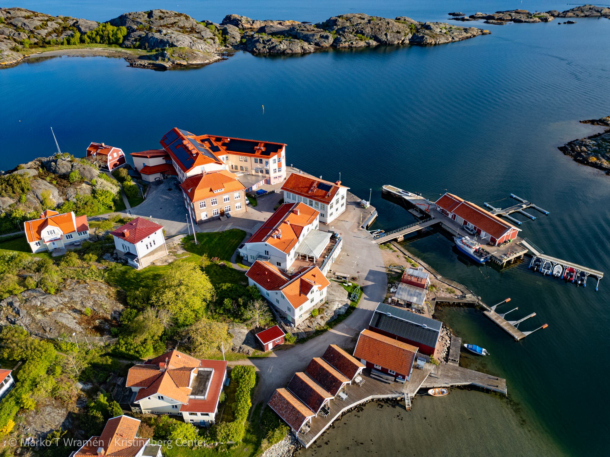 Luftaufnahme einer kleinen Küstensiedlung mit roten Dächern, umgeben von Wasser und felsigen Inseln. Mehrere Gebäude gruppieren sich um einen zentralen Platz, und ein Steg führt zu Booten im Wasser. Die Landschaft ist grün und felsig, mit klarem, blauem Wasser im Hintergrund.