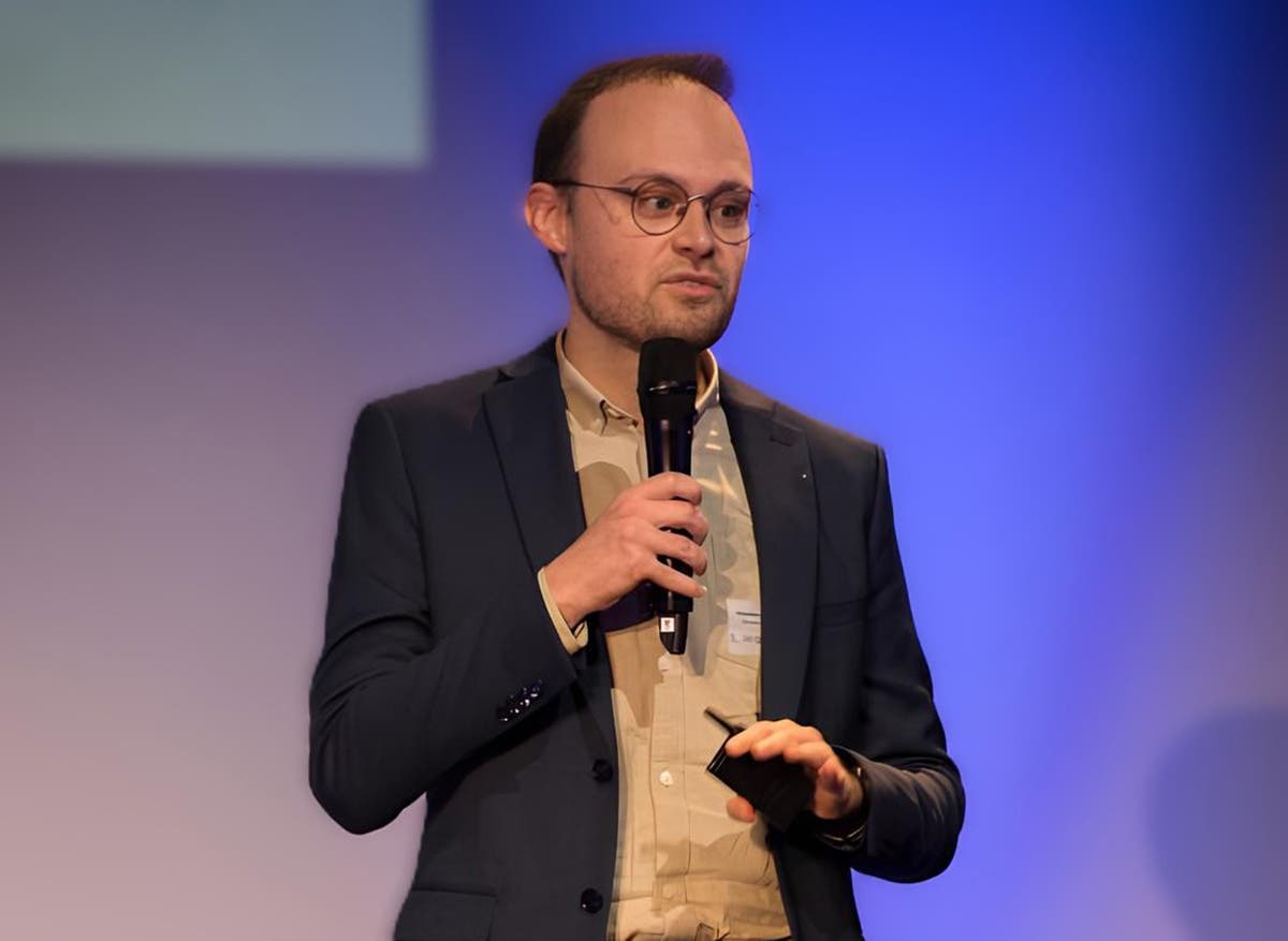 Eine Person in einem dunklen Anzug mit hellbraunem Hemd und Brille spricht auf einer Bühne mit einem Mikrofon in der rechten Hand. Die Person scheint eine Präsentation zu halten. Der Hintergrund ist in Blautönen gehalten. 