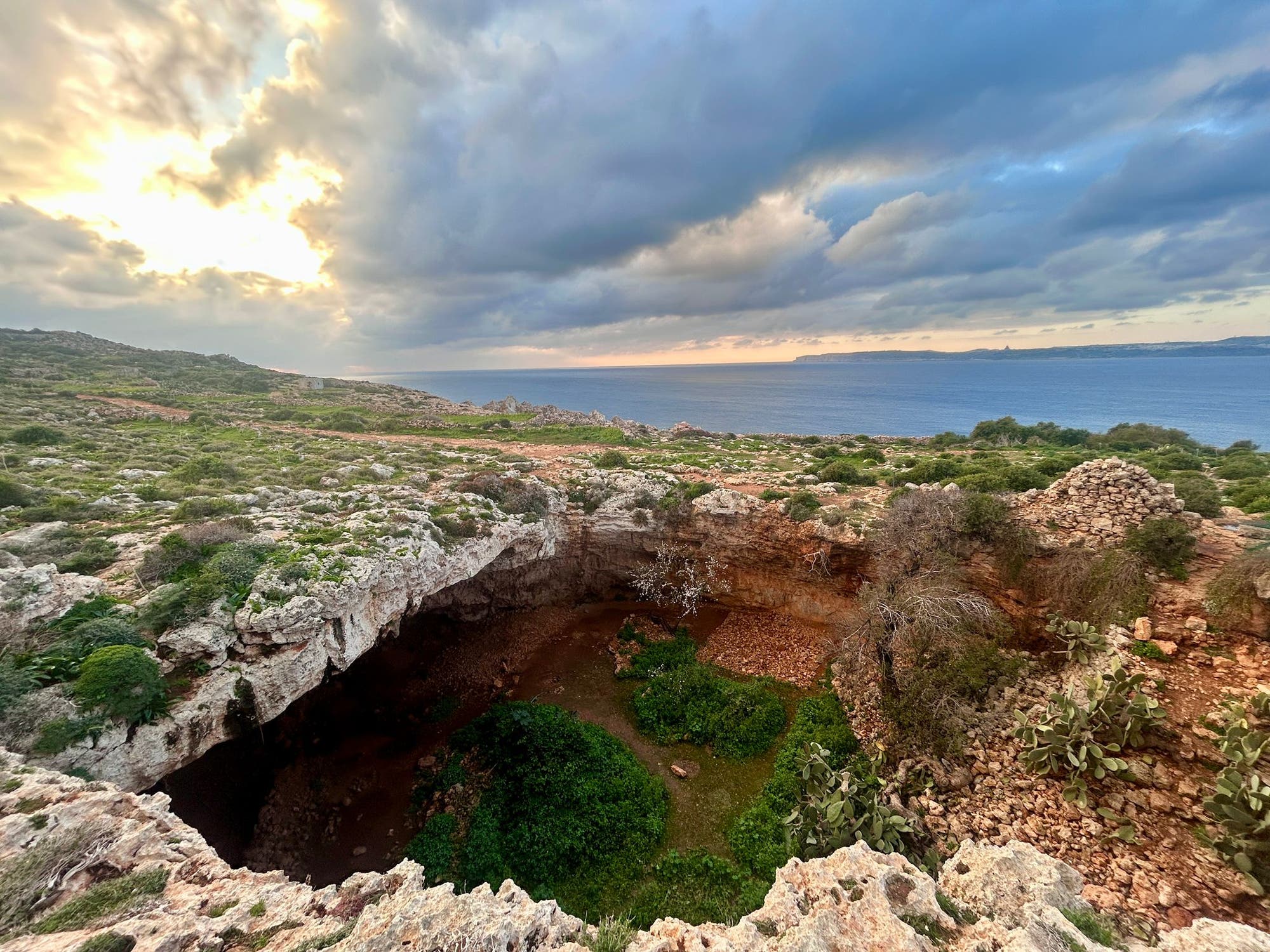 Eine malerische Küstenlandschaft mit einer großen, natürlichen Felsformation im Vordergrund, die eine grüne Senke umgibt. Der Himmel ist bewölkt, mit Sonnenstrahlen, die durch die Wolken brechen. Im Hintergrund erstreckt sich das blaue Meer bis zum Horizont. Die Umgebung ist von niedriger Vegetation und Felsen geprägt.