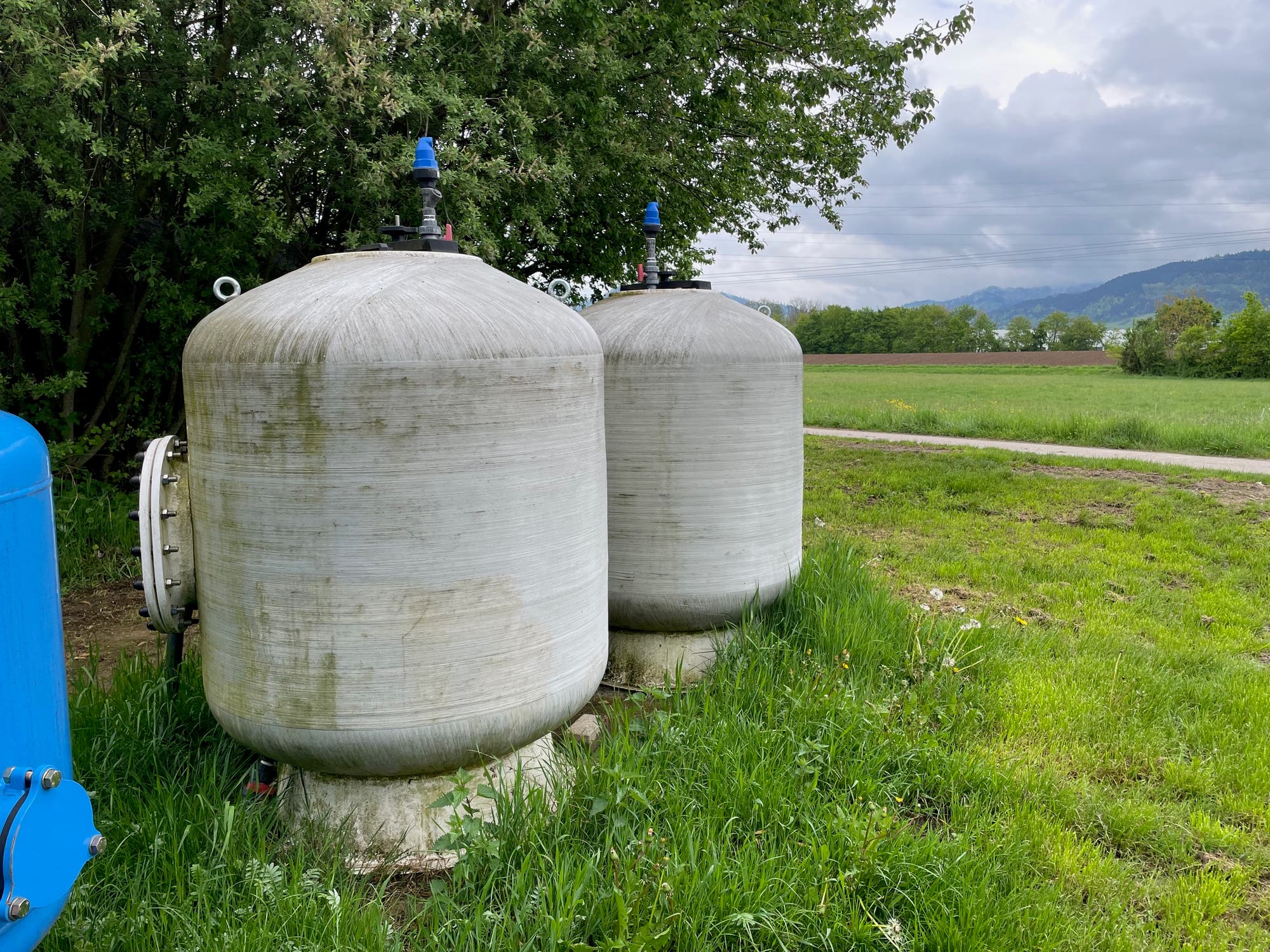 Zwei große, zylindrische Wassertanks aus Metall stehen nebeneinander auf einer grünen Wiese. Im Hintergrund sind Bäume und ein bewölkter Himmel zu sehen. Links im Bild befindet sich ein weiterer blauer Tank. Die Szene vermittelt einen ländlichen Eindruck.