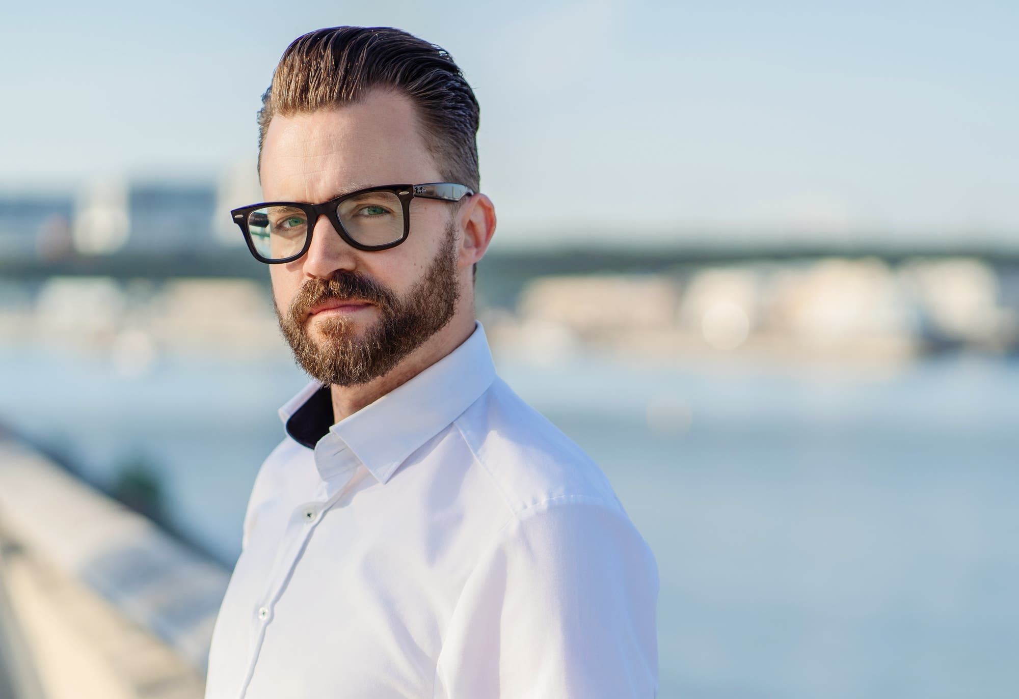 Ein Mann mit Brille und Bart steht in einem weißen Hemd am Wasser. Im Hintergrund ist eine verschwommene Stadtlandschaft mit einer Brücke zu sehen. Der Himmel ist klar und blau.