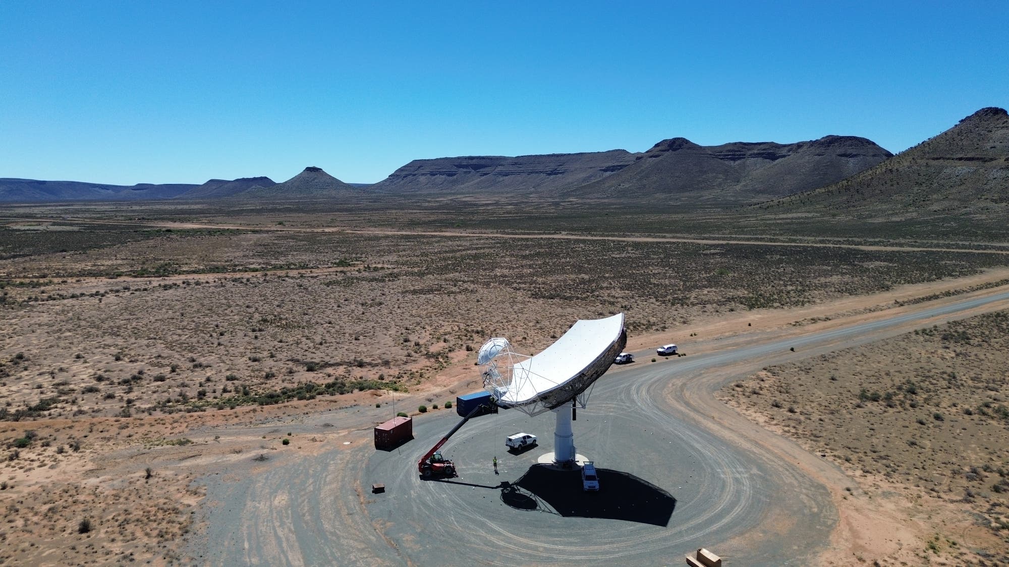 Eine große Satellitenschüssel steht in einer trockenen, weitläufigen Wüstenlandschaft mit Bergen im Hintergrund. Die Schüssel ist auf einem runden, unbefestigten Platz installiert, umgeben von einigen Fahrzeugen und Ausrüstungsgegenständen. Der Himmel ist klar und blau. Die Szene vermittelt ein Gefühl von Isolation und technologischer Erkundung in einer abgelegenen Umgebung.