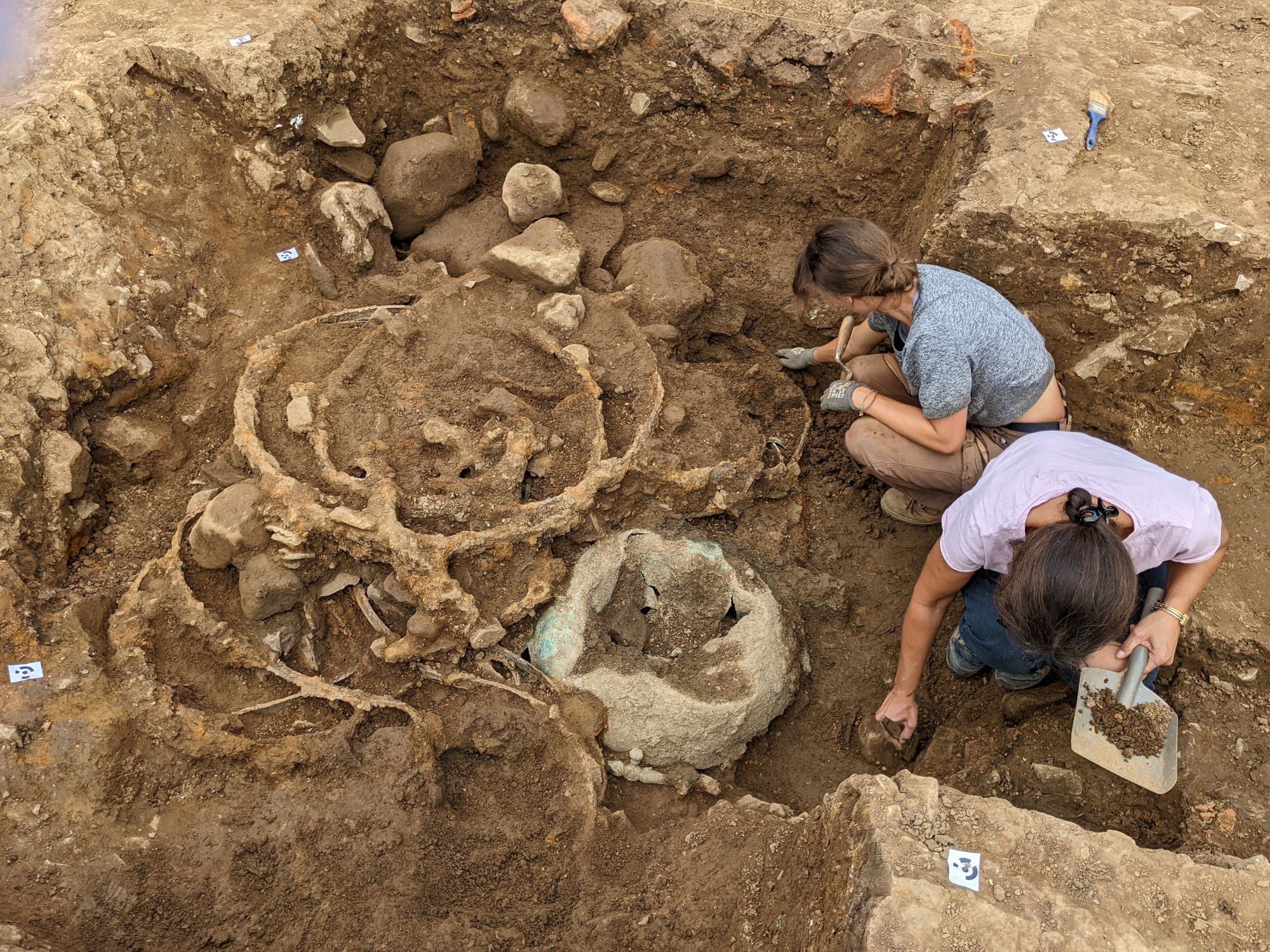 Zwei Archäologinnen arbeiten in einer Ausgrabungsstätte. Sie verwenden kleine Werkzeuge, um vorsichtig Erde um ein großes, kreisförmiges Artefakt zu entfernen, das von Steinen umgeben ist. Die Umgebung ist erdig und zeigt verschiedene Schichten des Bodens. Es sind keine weiteren Personen oder Objekte im Bild zu sehen.