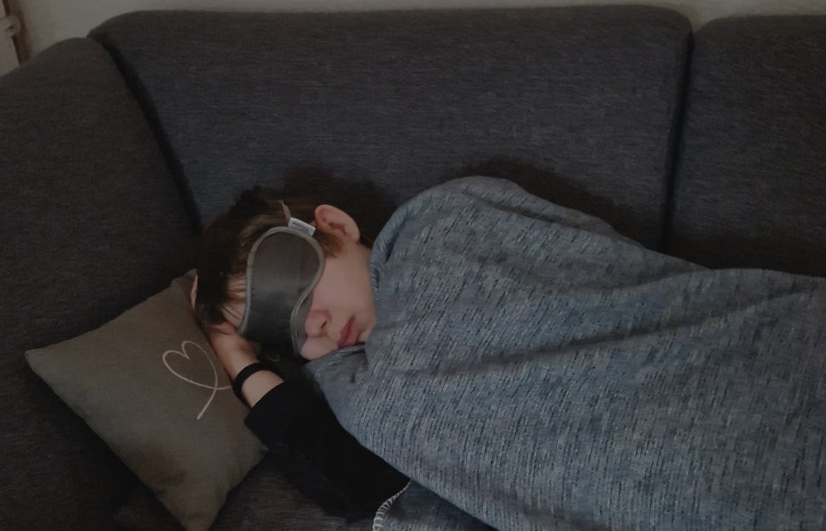 Eine Person liegt schlafend auf einem grauen Sofa, zugedeckt mit einer grauen Decke. Sie trägt eine Schlafmaske und ruht den Kopf auf einem Kissen mit einem aufgedruckten Herz. Die Szene vermittelt Ruhe und Entspannung.