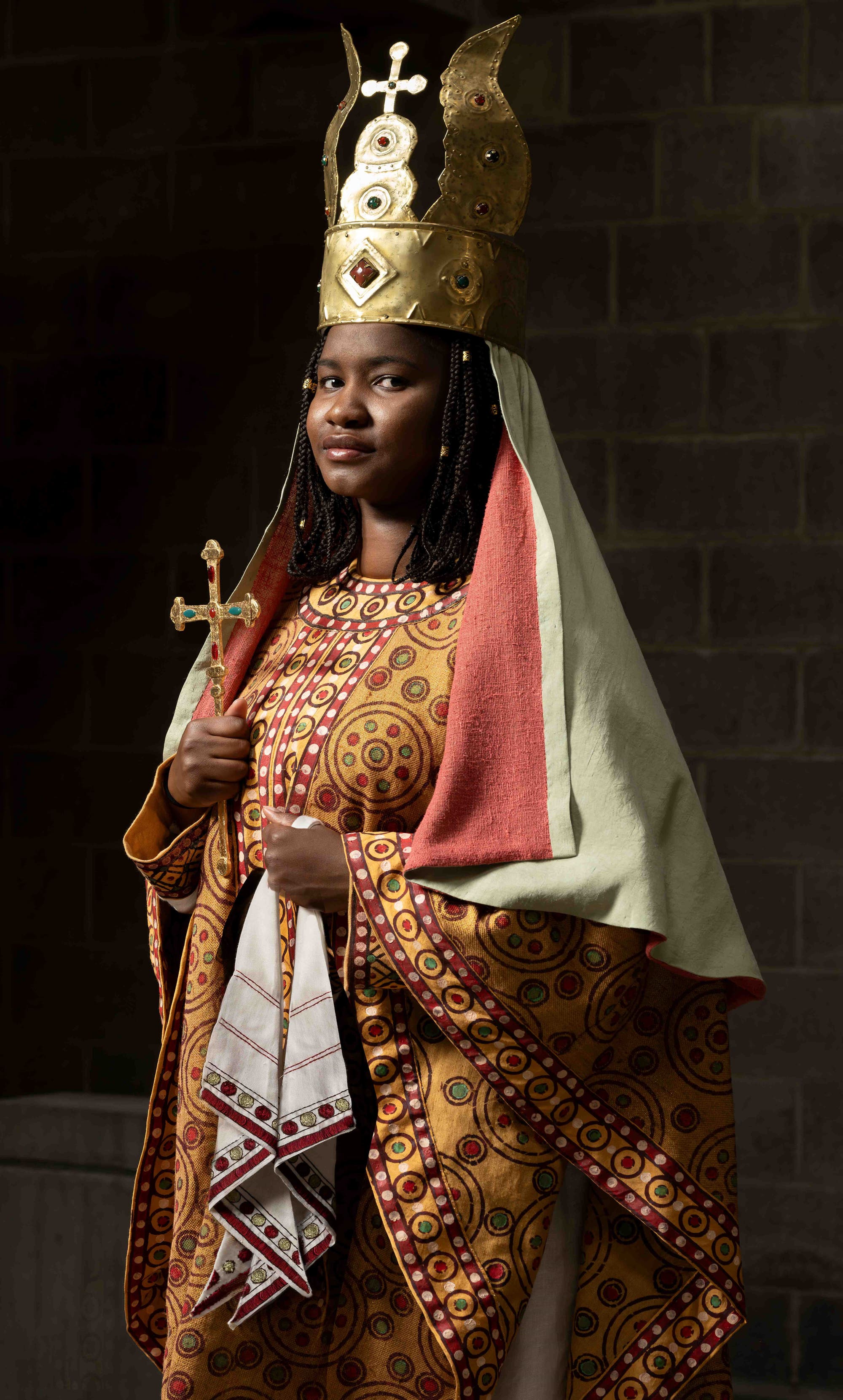 Eine Person in einem reich verzierten, traditionellen Gewand mit goldenen und roten Mustern steht vor einem dunklen Hintergrund. Sie trägt eine große, goldene Krone mit Verzierungen und hält ein Kreuz in der Hand. Ein rotes und cremefarbenes Tuch hängt über ihren Schultern. Die Person blickt selbstbewusst in die Kamera.
