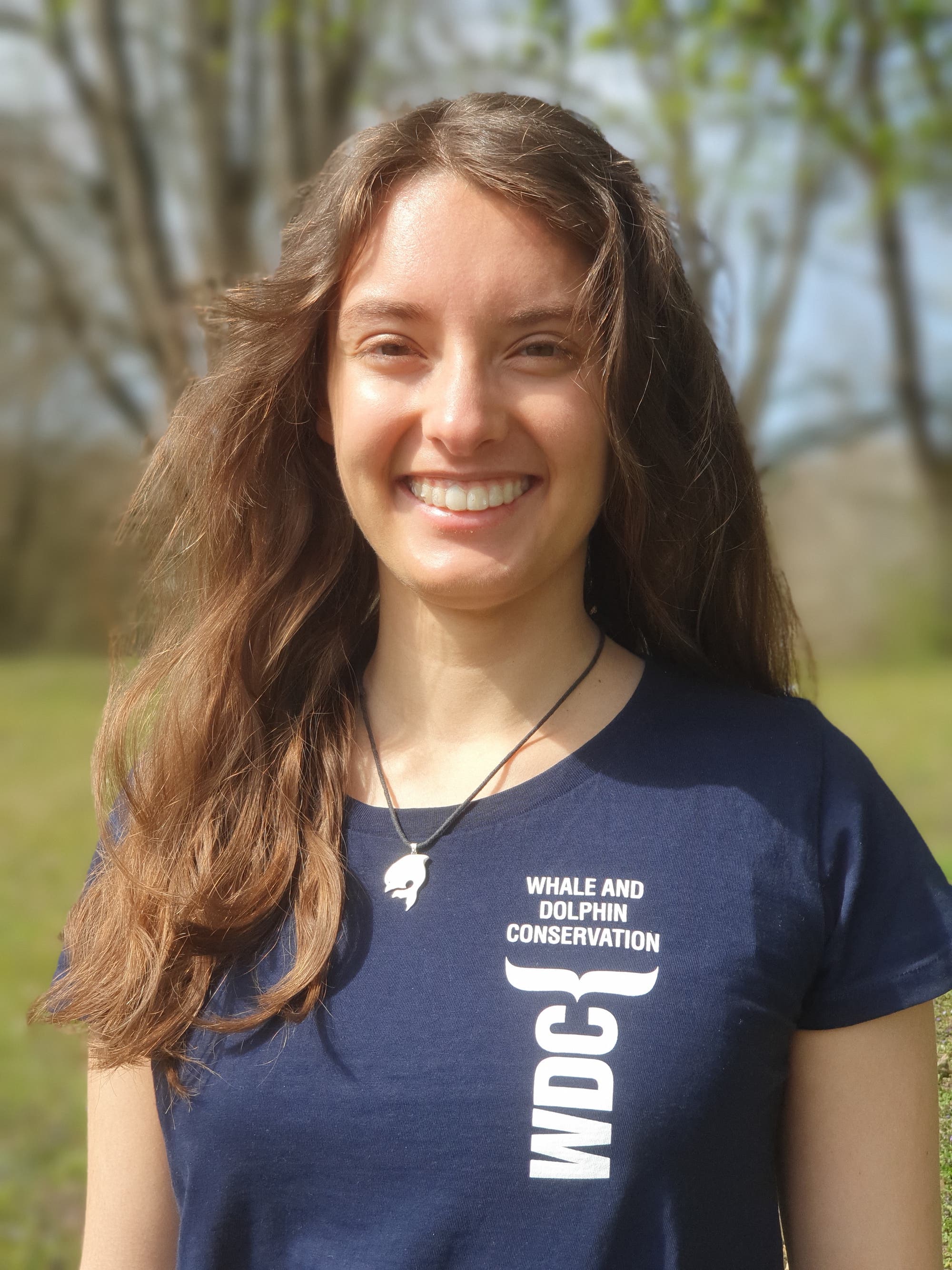 Eine Person steht im Freien vor einem See und lächelt in die Kamera. Sie trägt ein dunkelblaues T-Shirt mit der Aufschrift „Whale and Dolphin Conservation“. Im Hintergrund sind Bäume und ein klarer Himmel zu sehen.
