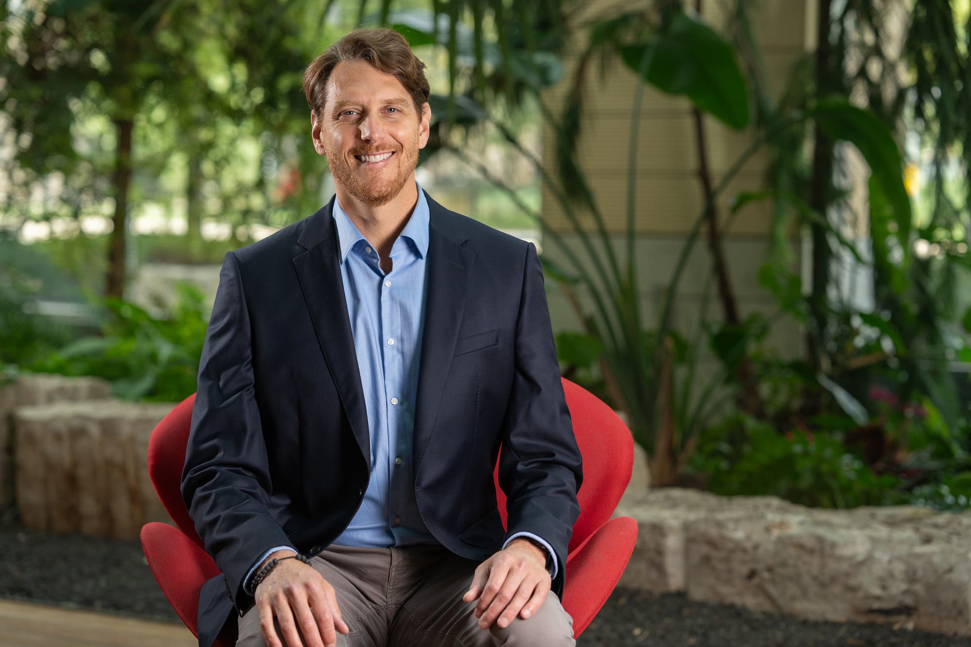 Ein Mann sitzt lächelnd auf einem roten Stuhl in einem Büro mit Pflanzen im Hintergrund. Er trägt ein dunkelblaues Sakko und ein hellblaues Hemd. 