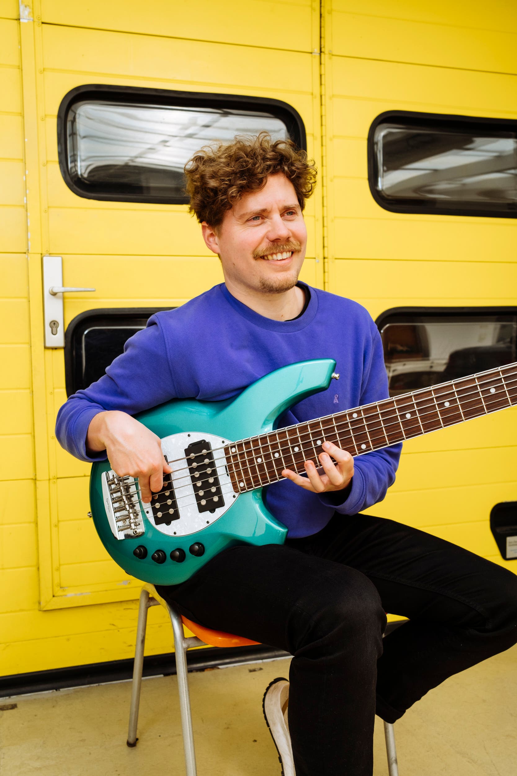 Ein Mann mit lockigem Haar sitzt auf einem Stuhl vor einem gelben Garagentor und spielt eine türkisfarbene E-Gitarre. Er trägt einen blauen Pullover und schwarze Hosen und lächelt, während er die Saiten zupft. Im Hintergrund ist eine weiße Tür mit einem Griff zu sehen.