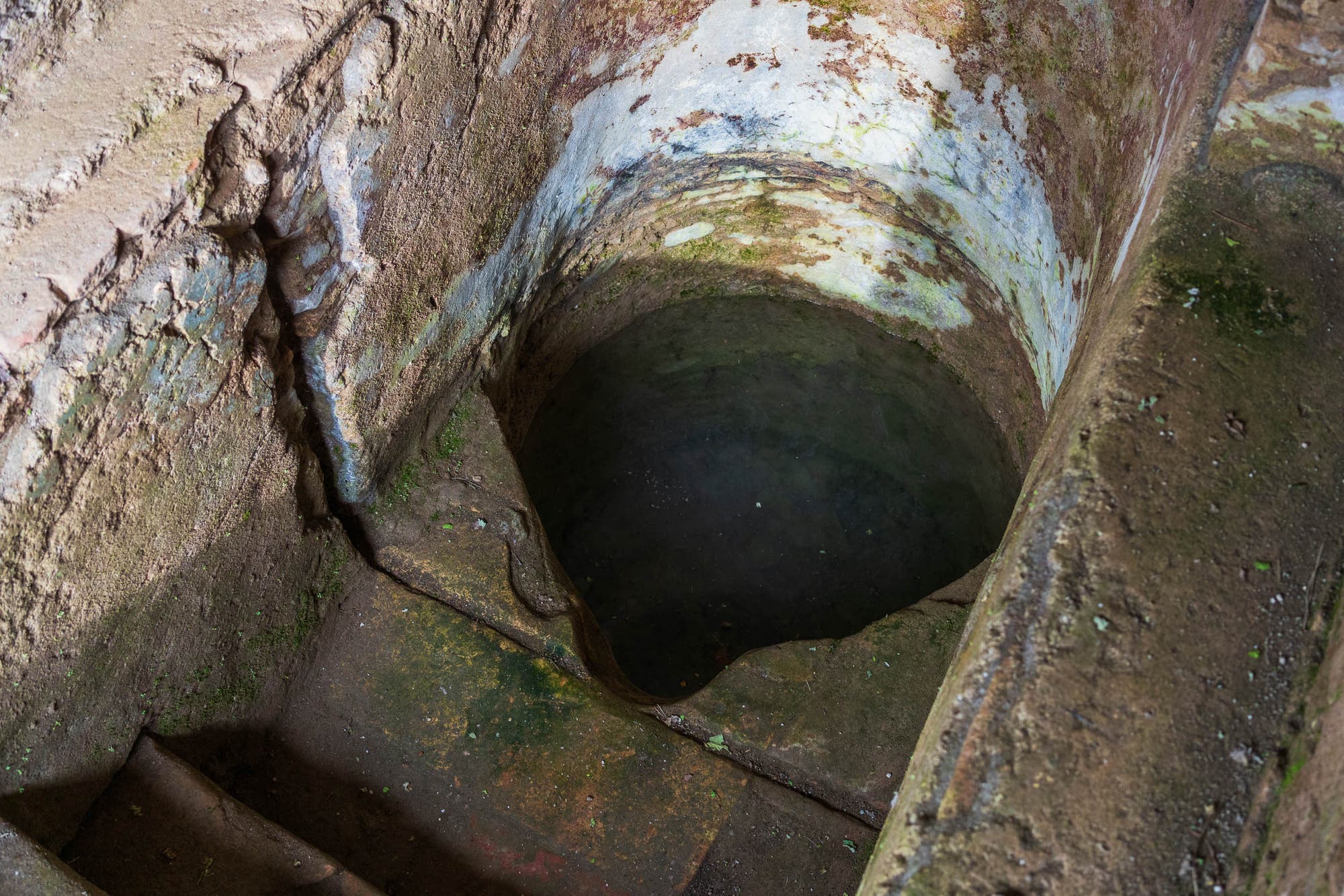 Die Öffnung einer spätantiker Mikwe mit moosbewachsenen Wänden und einer dunklen Wasseroberfläche am Boden. Die Struktur zeigt Abnutzungsspuren und Verfärbungen durch Feuchtigkeit. Die Öffnung befindet sich in einer Umgebung aus grobem Mauerwerk.