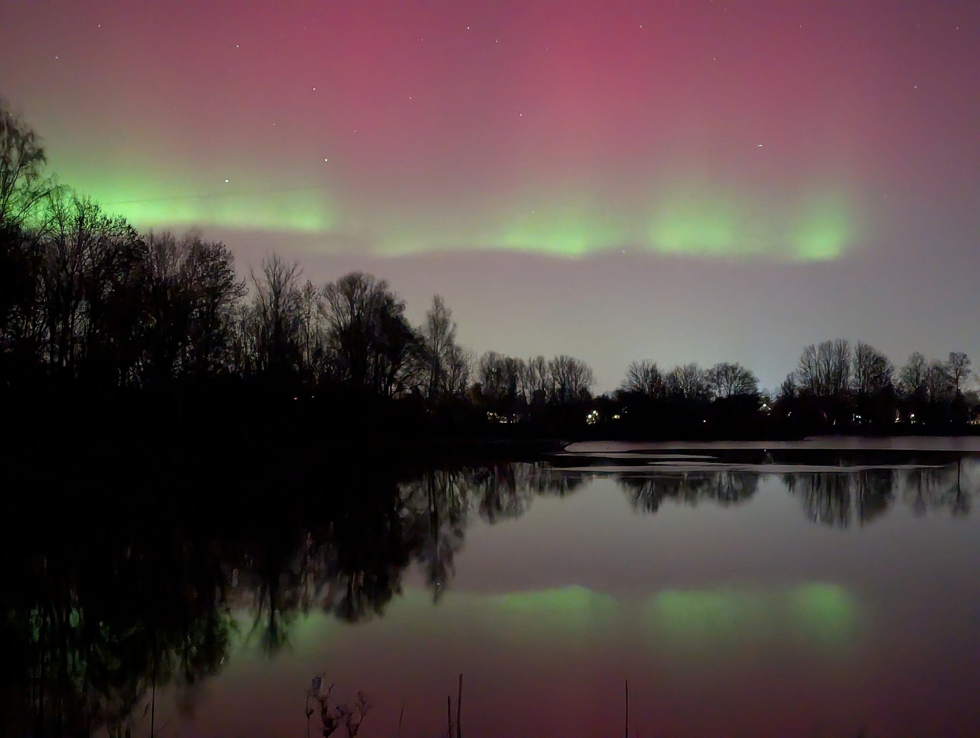 Eine n&auml;chtliche Landschaft mit einem See im Vordergrund, der die Silhouetten von B&auml;umen und das Nordlicht am Himmel reflektiert. Der Himmel zeigt gr&uuml;ne und rosa Farbt&ouml;ne, die sich &uuml;ber den Horizont erstrecken. Die Szene vermittelt eine ruhige und friedliche Atmosph&auml;re.