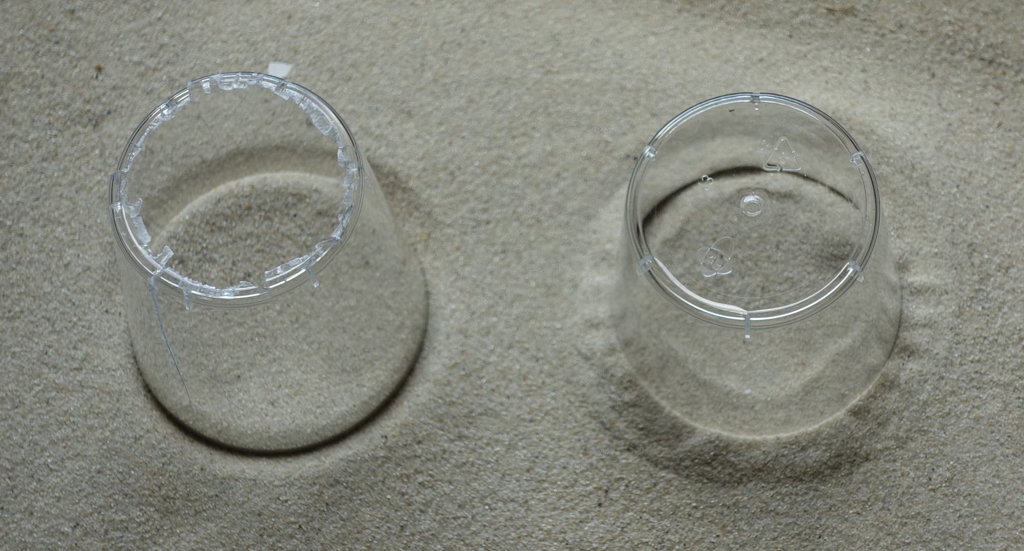 ein Plastikbecher ohne und ein Becher mit Boden sind nebeneinander in Sand gedrückt