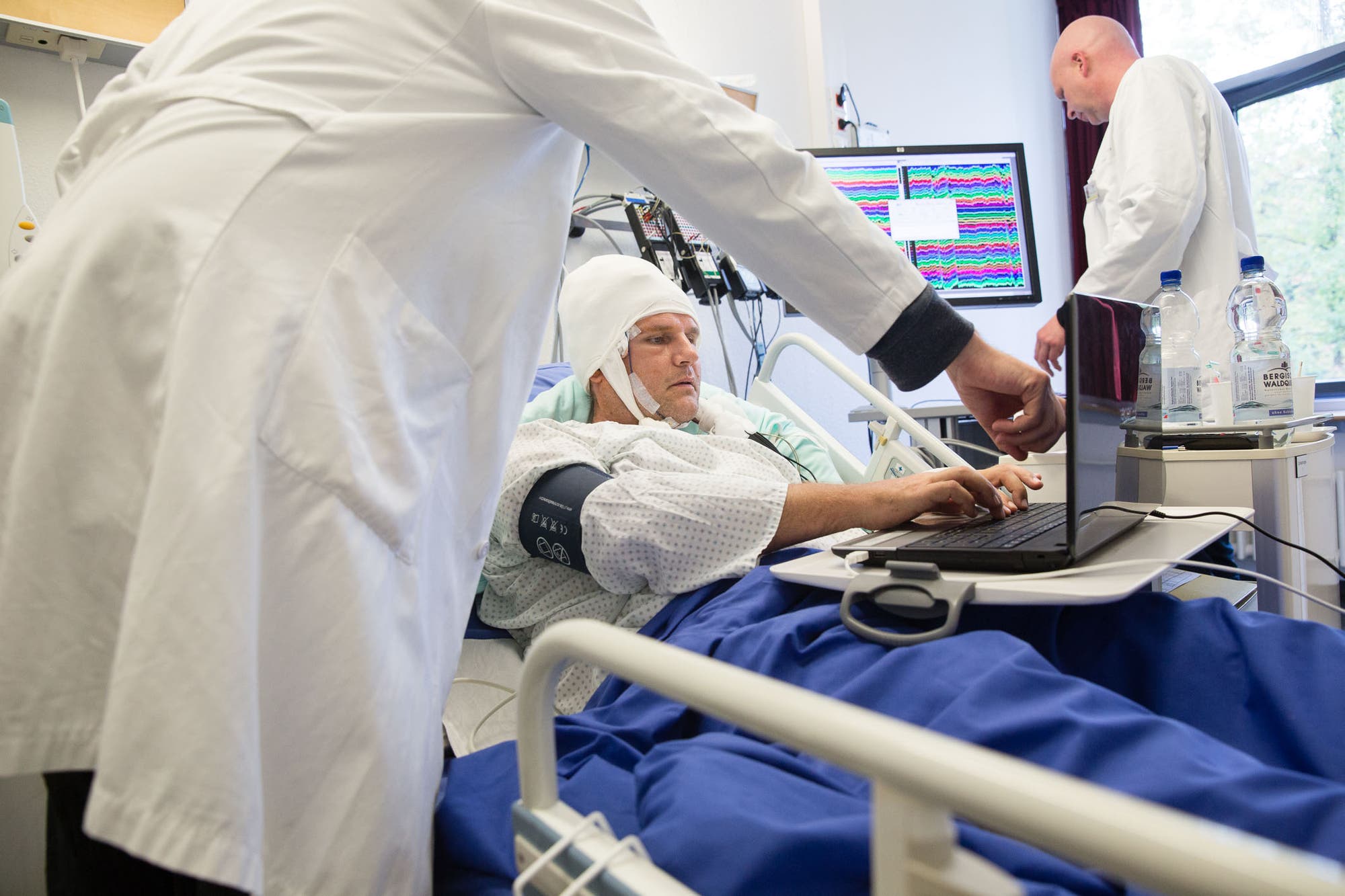 Ein Patient liegt im Krankenbett, vor ihm steht ein Laptop, auf den er, mit den Fingern auf der Tastatur, konzentriert schaut. Im Hintergrund sieht man einen Wissenschaftler, der sich die Hirnströme des Patienten ansieht