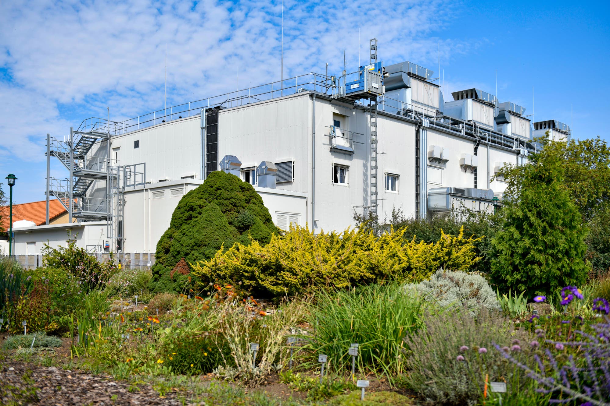 Ein modernes, weißes Gebäude mit mehreren Lüftungsanlagen und Treppen auf dem Dach, umgeben von einem üppigen Garten mit verschiedenen Pflanzenarten. Der Himmel ist leicht bewölkt. Im Vordergrund sind bunte Blumen und Sträucher zu sehen, die dem Bild eine lebendige Atmosphäre verleihen.