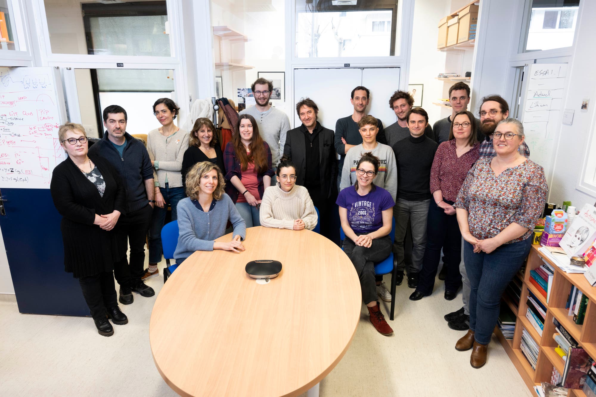 Eine Gruppe von 17 Personen steht und sitzt um einen ovalen Tisch in einem Büro. Die Gruppe gehört zum Team "Génétique humaine et fonctions cognitives" am Institut Pasteur in Paris, geleitet von Thomas Bourgeron. Sie forschen zu Genen, die mit Autismus und Gehirnfunktionen in Verbindung stehen. Im Hintergrund sind Whiteboards und Bücherregale zu sehen. Die Atmosphäre wirkt professionell und engagiert.