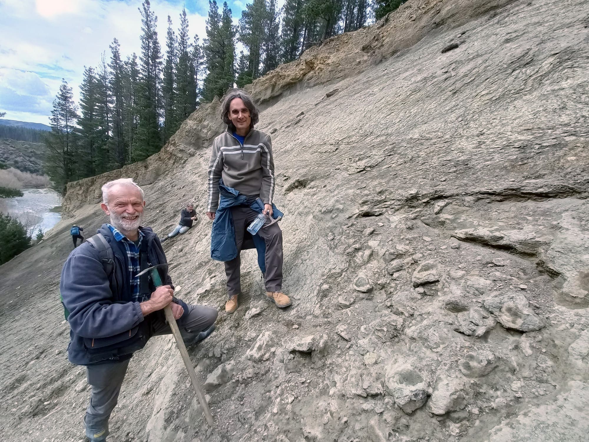 Zwei Personen stehen an einem steilen, felsigen Hang in einer bewaldeten Gegend. Der ältere Mann (links) hält eine Spitzhacke und lächelt, während der andere (rechts) eine Wasserflasche hält. Im Hintergrund sind Bäume und ein Fluss zu sehen. Die Szene vermittelt eine Outdoor-Aktivität in der Natur.