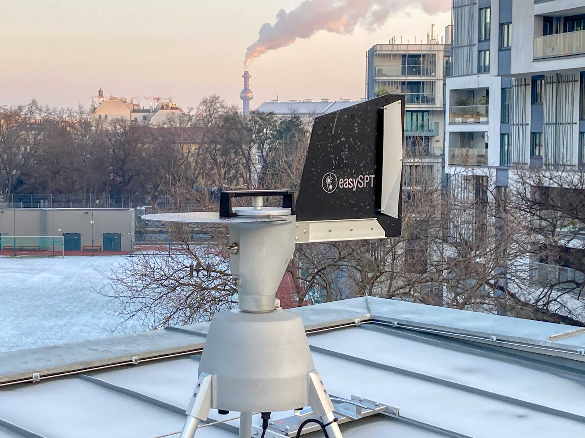 Ein Gerät mit der Aufschrift "easySPT" ist auf einem Dach montiert. Im Hintergrund sind moderne Gebäude und ein Schornstein zu sehen, aus dem Rauch aufsteigt. Die Umgebung ist winterlich mit Schnee bedeckt.