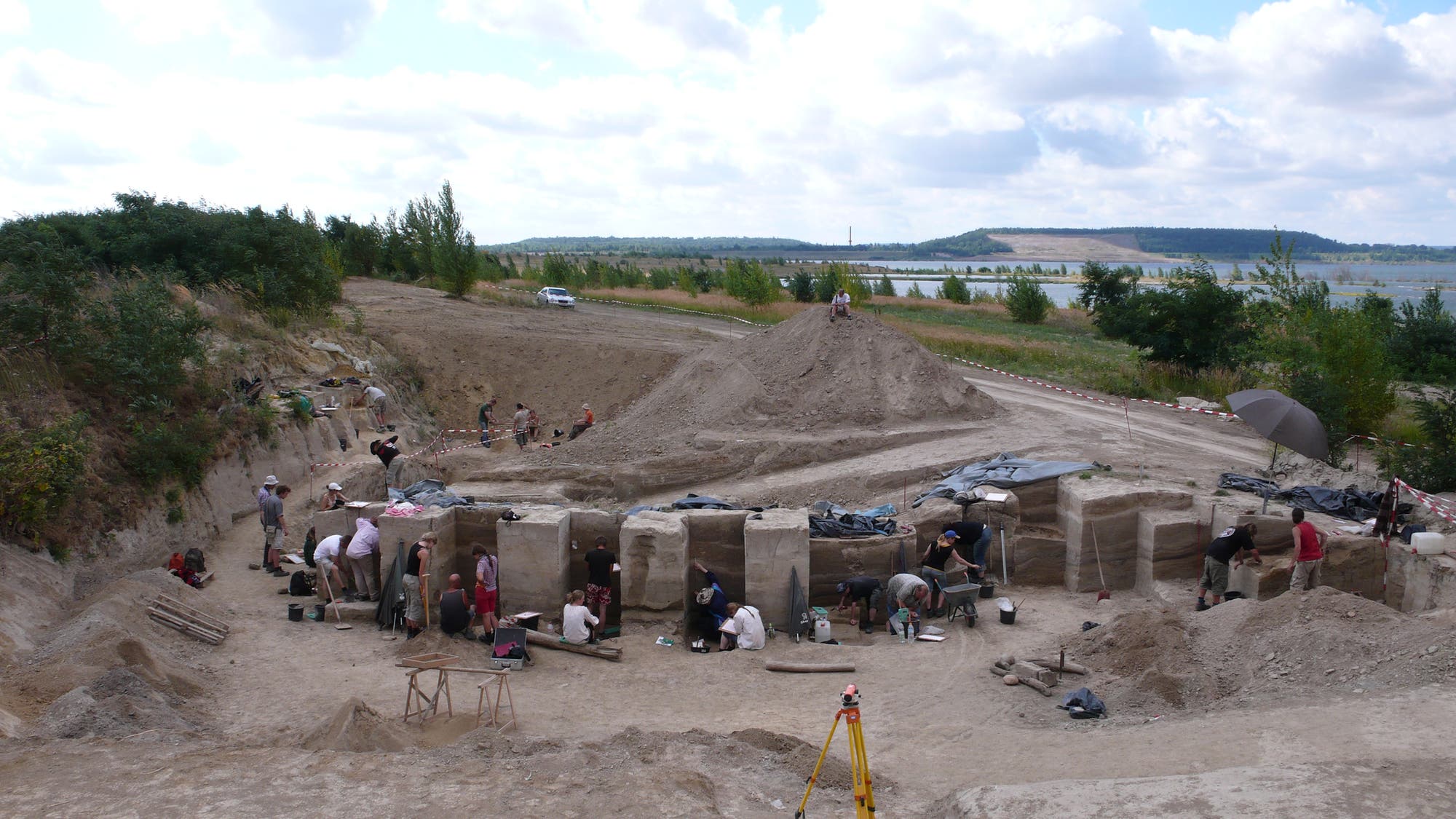 Eine archäologische Ausgrabungsstätte mit mehreren Menschen, die an verschiedenen Stellen arbeiten. Sie sind mit Werkzeugen beschäftigt, um Erde und Artefakte freizulegen. Im Vordergrund sind große, rechteckige Erdblöcke zu sehen, die sorgfältig freigelegt wurden. Ein Stativ steht im Sand, möglicherweise für Vermessungszwecke. Im Hintergrund erstreckt sich eine hügelige Landschaft mit Bäumen und einem See unter einem bewölkten Himmel.