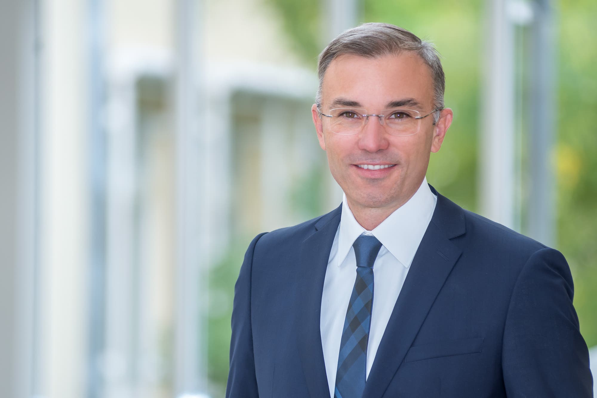 Eine Person in einem Anzug steht in einem hellen, modernen Innenraum mit großen Fenstern. Die Person lächelt leicht und trägt eine Brille. Im Hintergrund sind verschwommen grüne Pflanzen zu sehen, die dem Raum eine freundliche Atmosphäre verleihen.