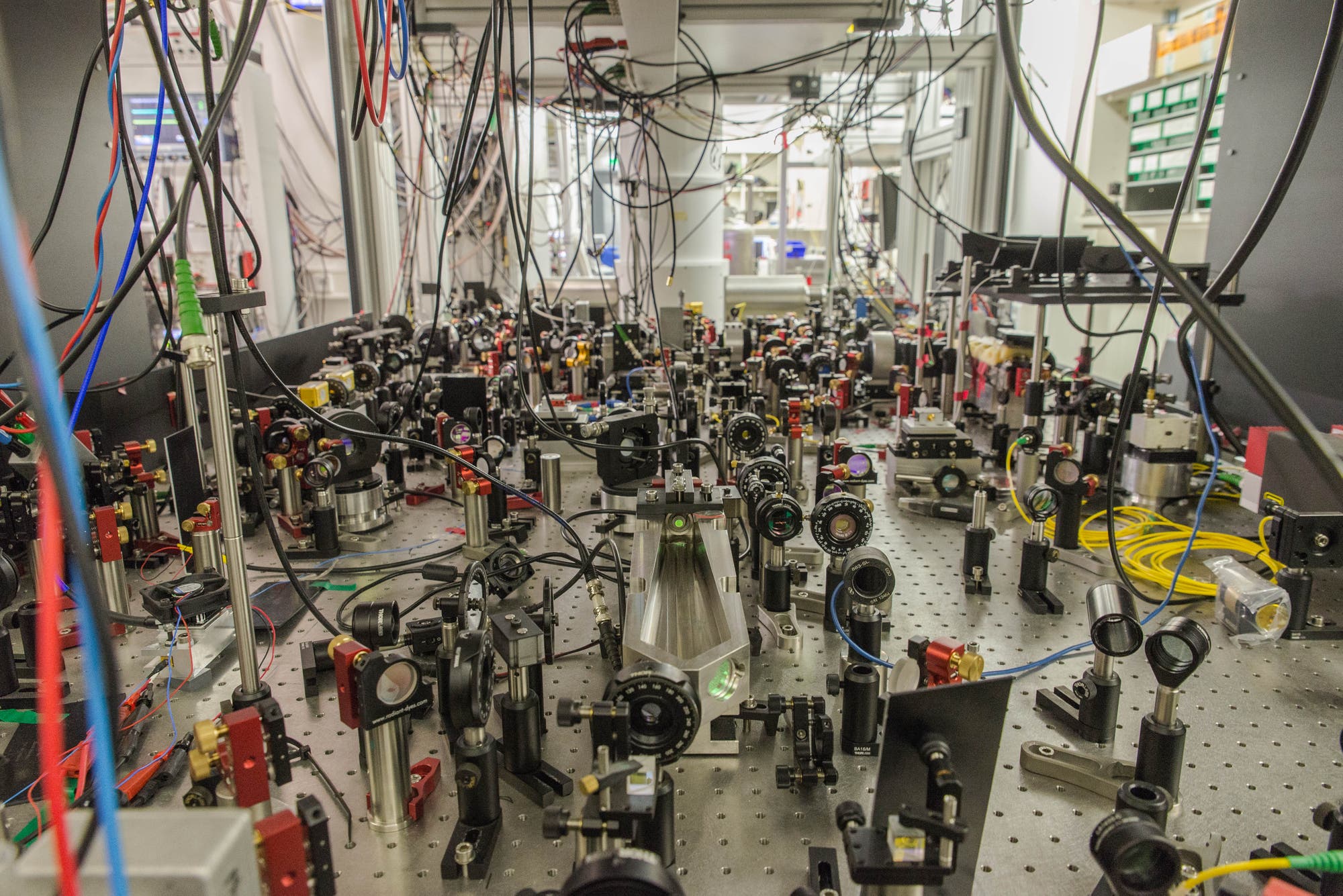Ein Labor mit einer komplexen Anordnung von optischen Geräten und Lasern auf einem optischen Tisch. Zahlreiche Kabel und Drähte verlaufen über die Geräte, die aus Linsen, Spiegeln und Halterungen bestehen. Im Hintergrund sind weitere technische Geräte und Regale zu sehen. Die Szene vermittelt den Eindruck eines hochspezialisierten wissenschaftlichen Experiments.