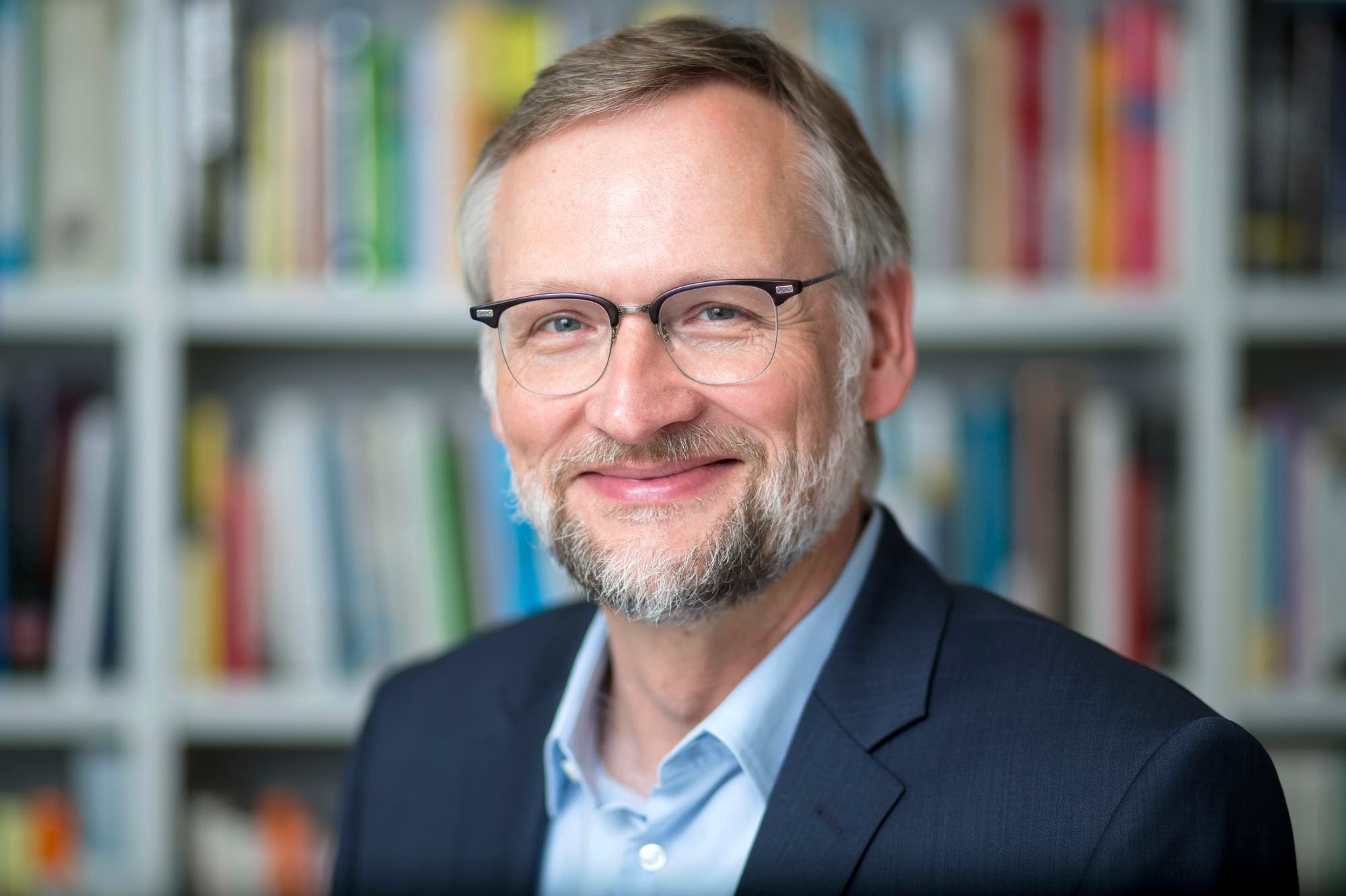 Ein lächelnder Mann mit Brille und Bart in einem Anzug ist vor einem Bücherregal zu sehen. Der Hintergrund ist unscharf, so dass die vielen Bücher nicht klar erkennbar sind.