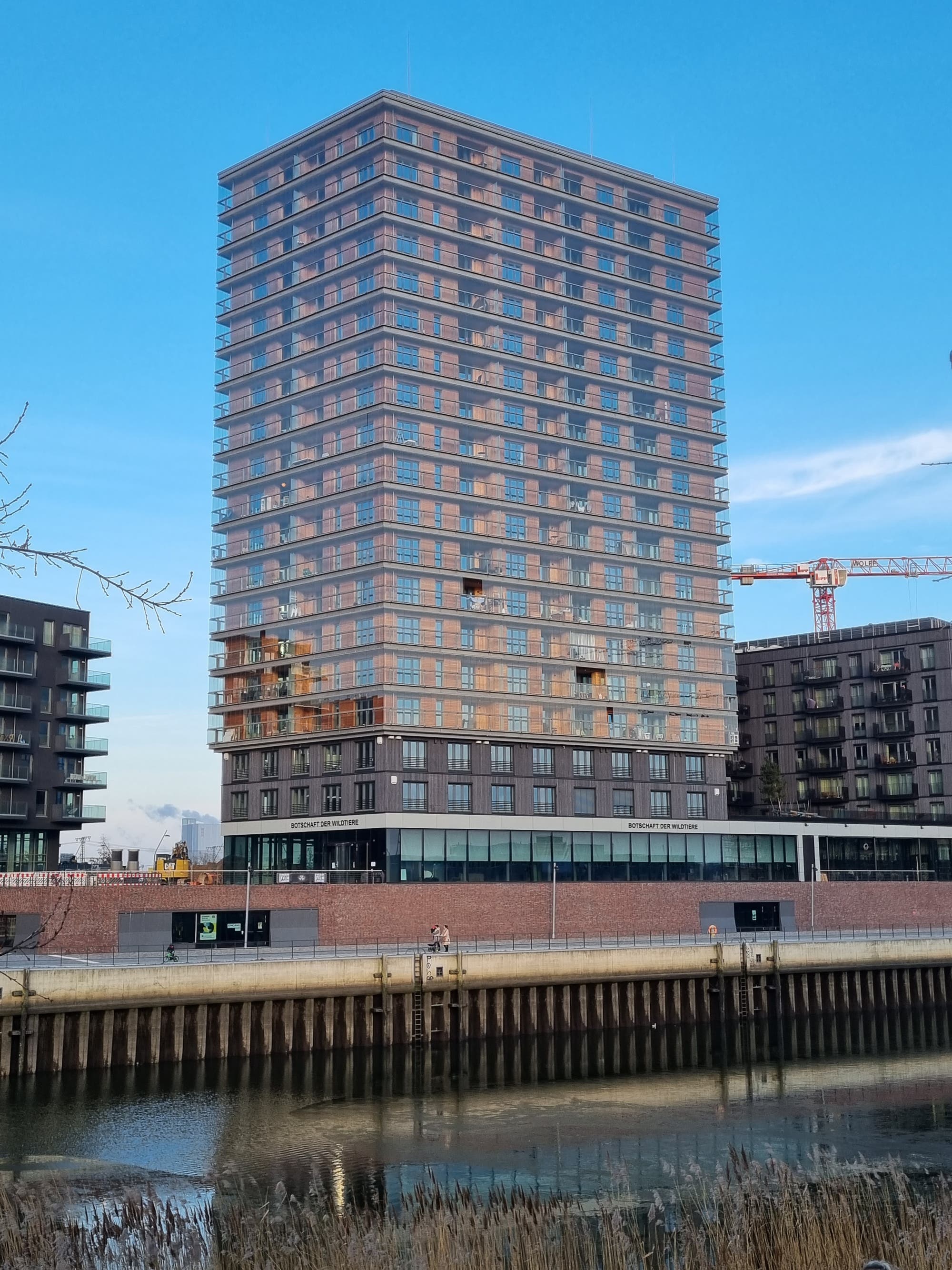 Ein modernes Hochhaus mit vielen Fenstern spiegelt sich im Wasser eines Kanals wider. Das Gebäude hat eine Fassade aus Glas und Holz und zeigt, dass man mit Holz genauso hässlich bauen kann wie mit Stahlbeton. 