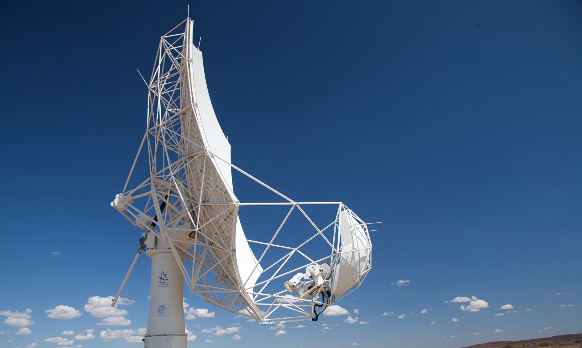 Eine große, weiße Satellitenschüssel steht vor einem klaren blauen Himmel. Die Struktur ist komplex und besteht aus einem Netzwerk von Metallstreben. Im Hintergrund sind einige weiße Wolken zu sehen. Die Schüssel ist auf einem stabilen Sockel montiert, der mit Logos versehen ist. Es gibt keine Menschen im Bild. Die Szene vermittelt ein Gefühl von Technologie und Kommunikation.