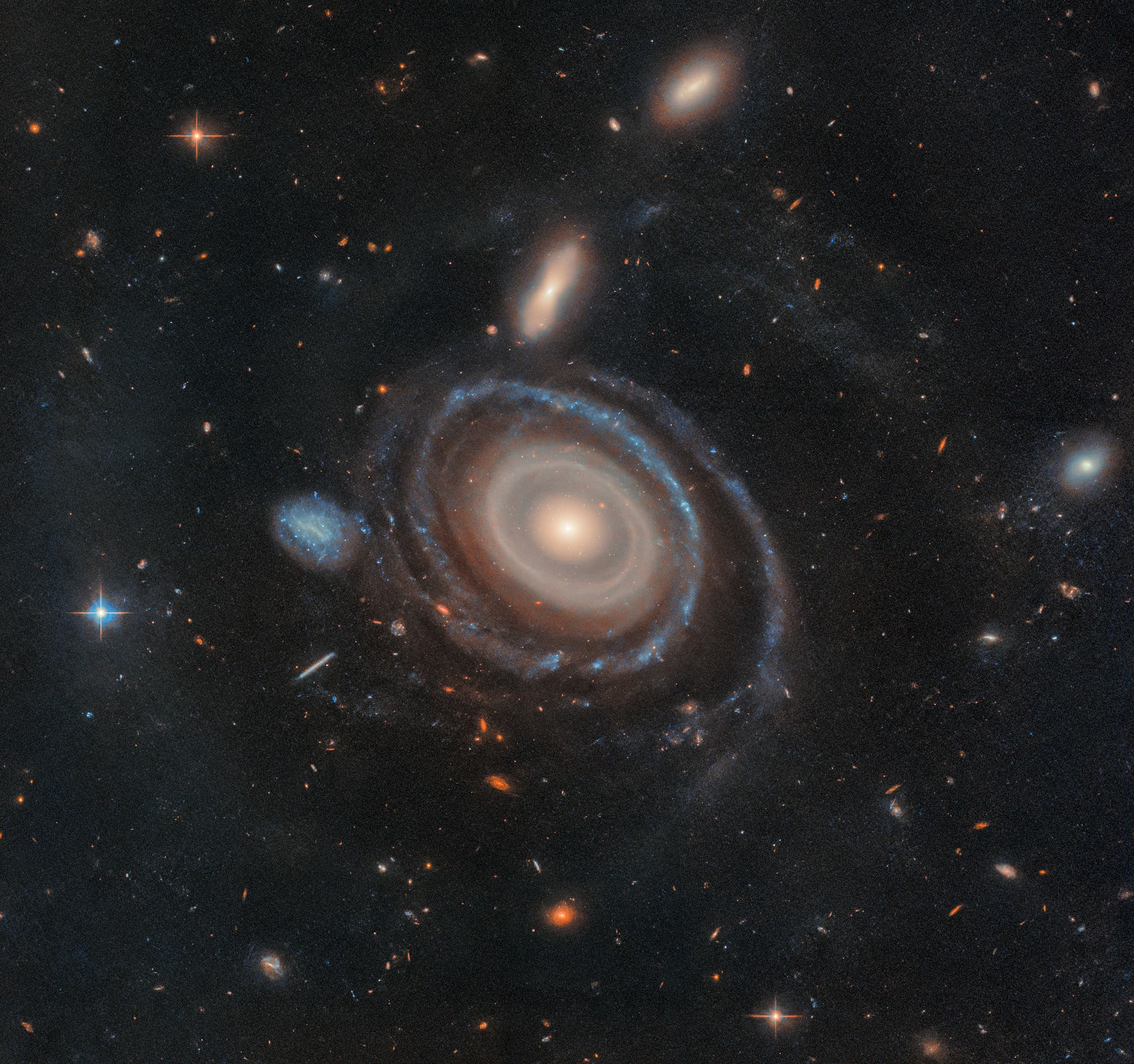 Eine Spiralgalaxie mit mehreren leuchtenden Armen, die sich um einen hellen Kern winden, ist im Zentrum des Bildes zu sehen. Sie ist von zahlreichen kleineren Galaxien und Sternen umgeben, die im dunklen Weltraum verstreut sind. Die Galaxie zeigt eine klare Struktur mit bläulichen und rötlichen Farbtönen.