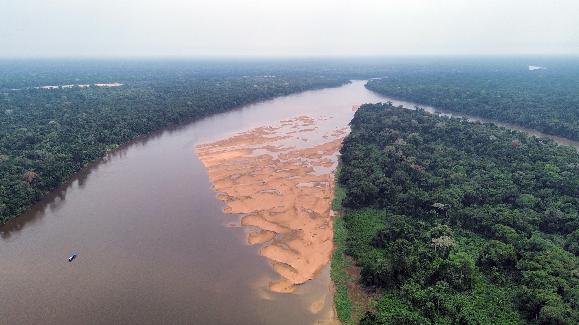 Luftaufnahme eines breiten Flusses, der durch einen dichten, grünen Regenwald fließt. Auf der rechten Seite des Flusses befindet sich eine große Sandbank. Der Himmel ist bewölkt, und am linken Rand des Bildes ist ein kleines Boot auf dem Wasser zu sehen. Die Szene vermittelt die natürliche Schönheit und Abgeschiedenheit der Landschaft.