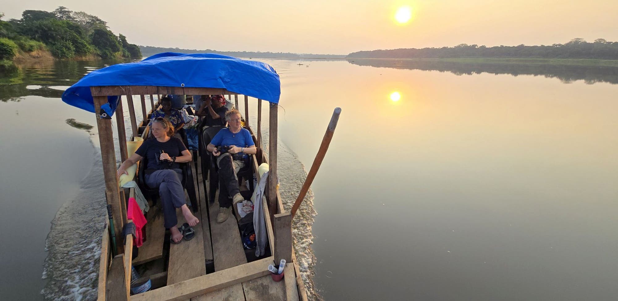 Eine Gruppe von Menschen sitzt entspannt in einem kleinen, überdachten Holzboot, das einen ruhigen Fluss entlangfährt. Die Sonne steht tief am Himmel und spiegelt sich im Wasser, was eine friedliche Stimmung erzeugt. Im Hintergrund sind dichte Bäume am Flussufer zu sehen.