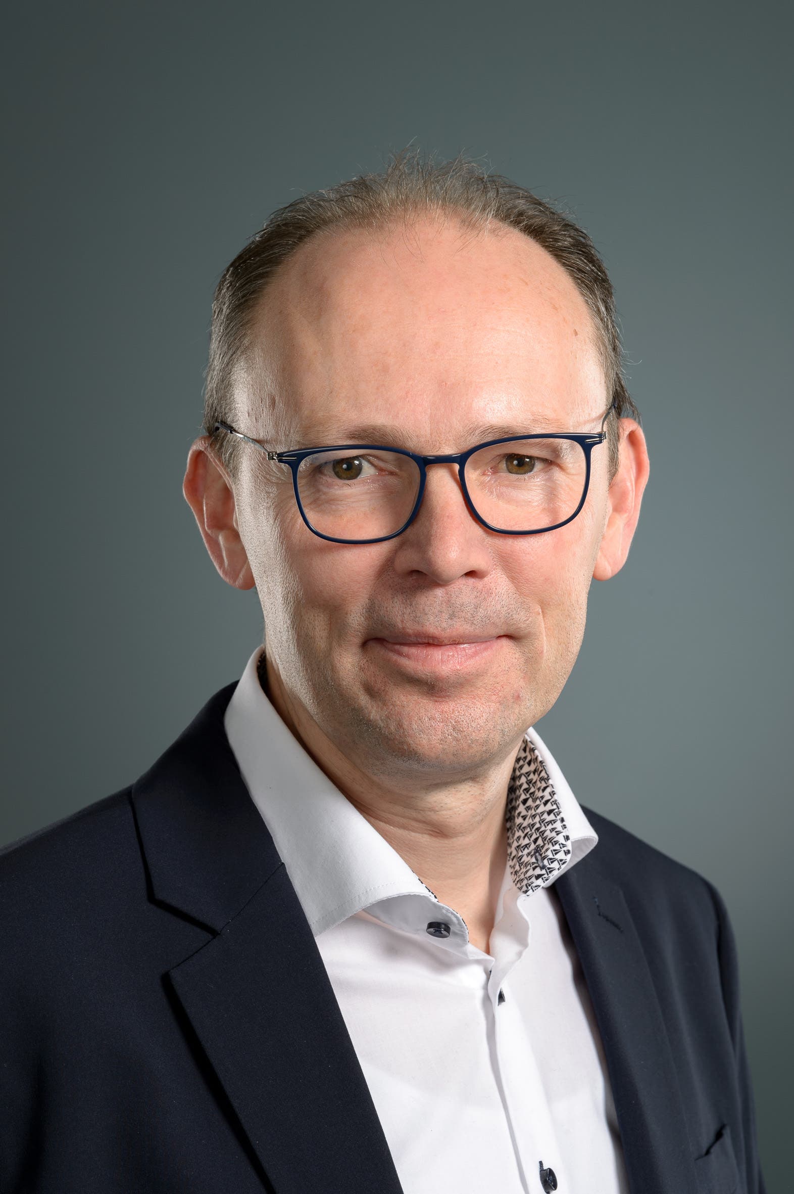 Eine Person in einem formellen Anzug mit Brille lächelt leicht in die Kamera. Der Hintergrund ist neutral grau, was den Fokus auf die Person lenkt. Die Person trägt ein weißes Hemd mit einem gemusterten Kragen.