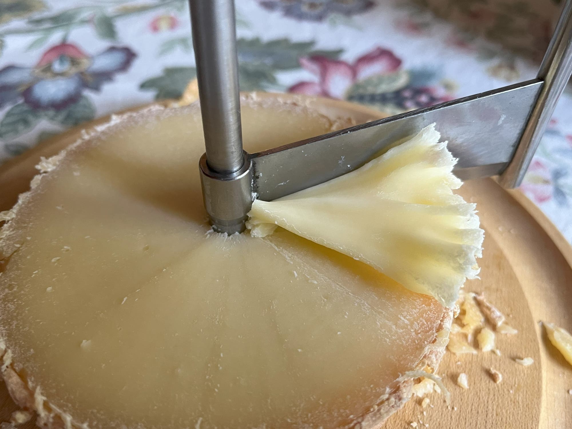 Ein Tête-de-Moine-Käse wird mit einem Girolle-Käsehobel geschnitten. Der Hobel schabt eine dünne, dekorative Rosette vom Käse ab. Der Käse liegt auf einem hölzernen Brett, im Hintergrund ist ein Tisch mit blumigem Tischtuch zu sehen.