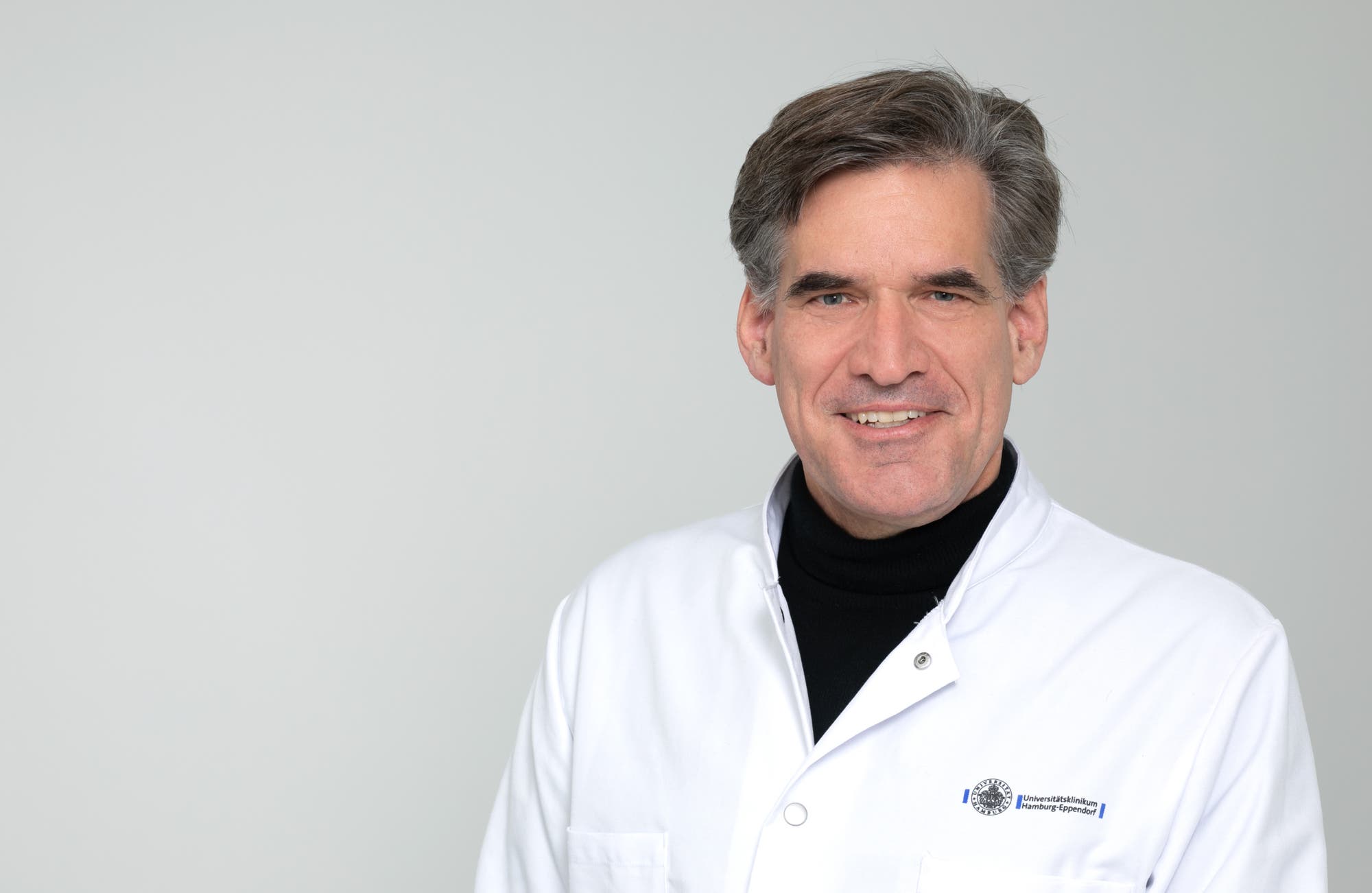 Eine Person in einem weißen Arztkittel mit einem Logo der Universitätsklinikum Hamburg-Eppendorf. Die Person lächelt und blickt in die Kamera. Der Hintergrund ist neutral und hell.