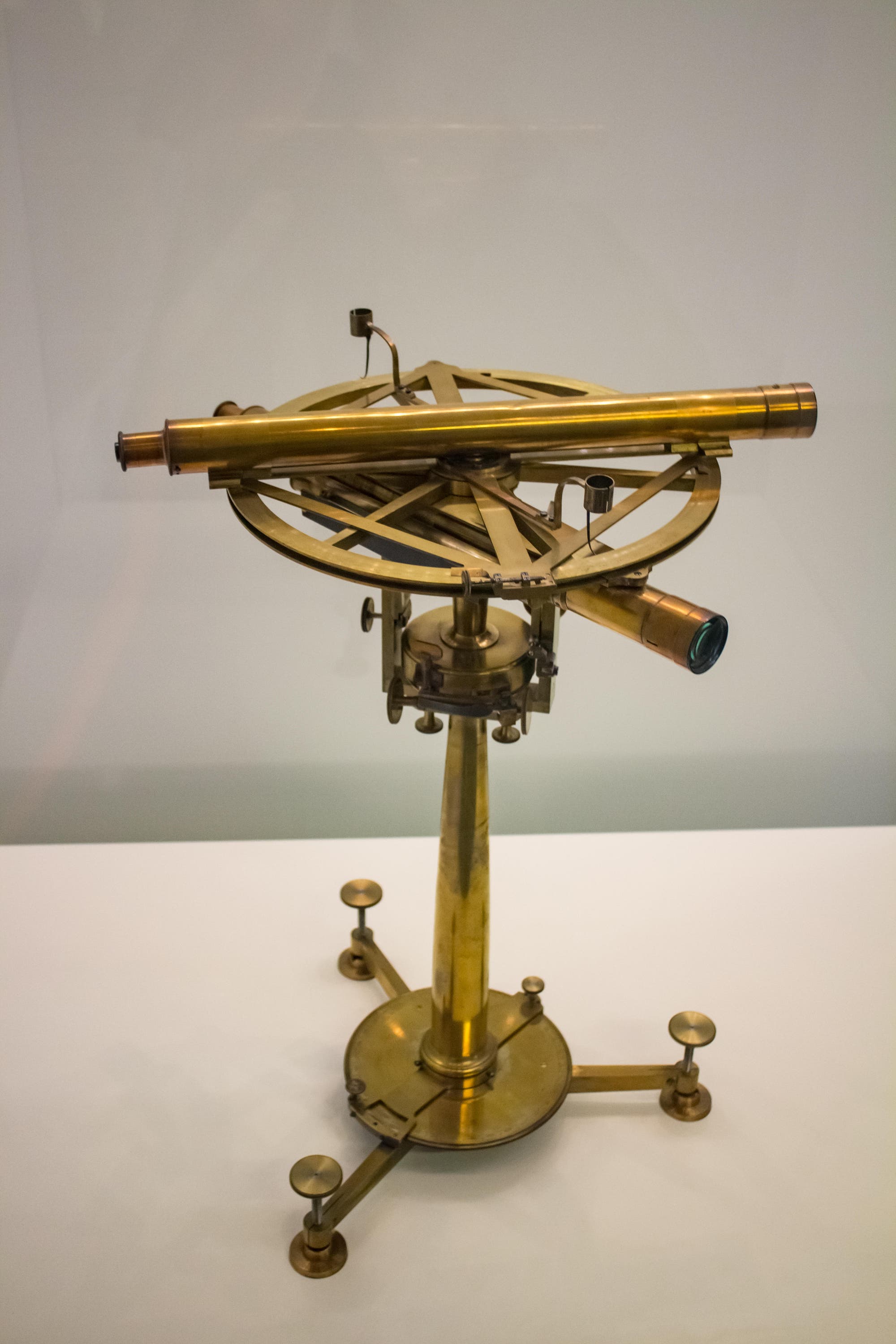 Ein antikes, aus Messing gefertigtes Theodolit-Instrument steht auf einem weißen Tisch. Es verfügt über ein Teleskop, das auf einem drehbaren Kreis montiert ist, und ist mit mehreren Einstellschrauben ausgestattet. Das Gerät wird von einem stabilen Stativ getragen. Im Hintergrund ist eine neutrale, graue Wand zu sehen. 