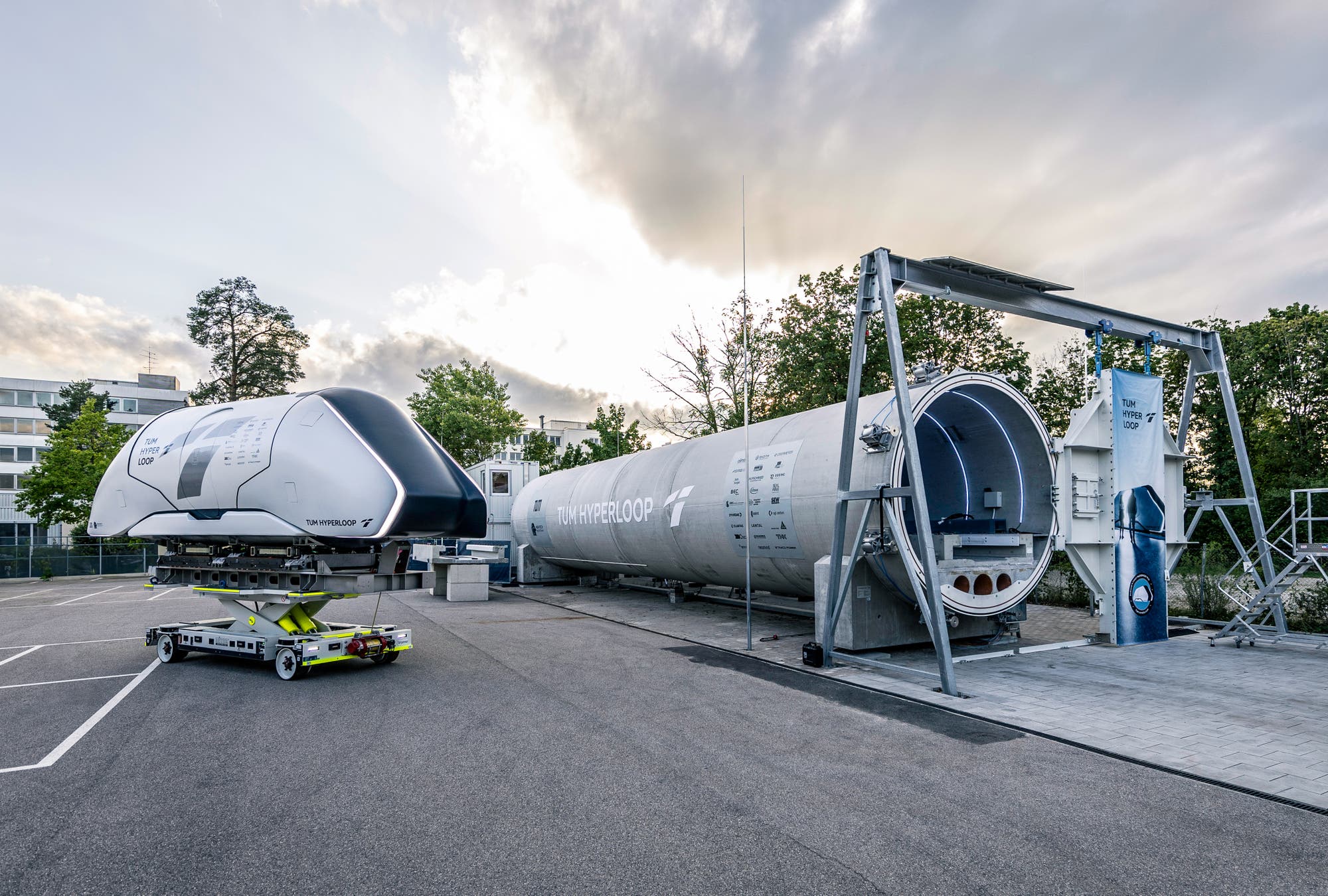 Eine Hyperloop-Testanlage im Freien bei bewölktem Himmel. Links ist eine futuristische Transportkapsel auf einem mobilen Träger zu sehen, rechts eine lange, zylindrische Röhre mit der Aufschrift "TUM Hyperloop". Die Umgebung ist von Bäumen und Gebäuden umgeben. Die Szene vermittelt den technologischen Fortschritt im Bereich Hochgeschwindigkeitsverkehr.