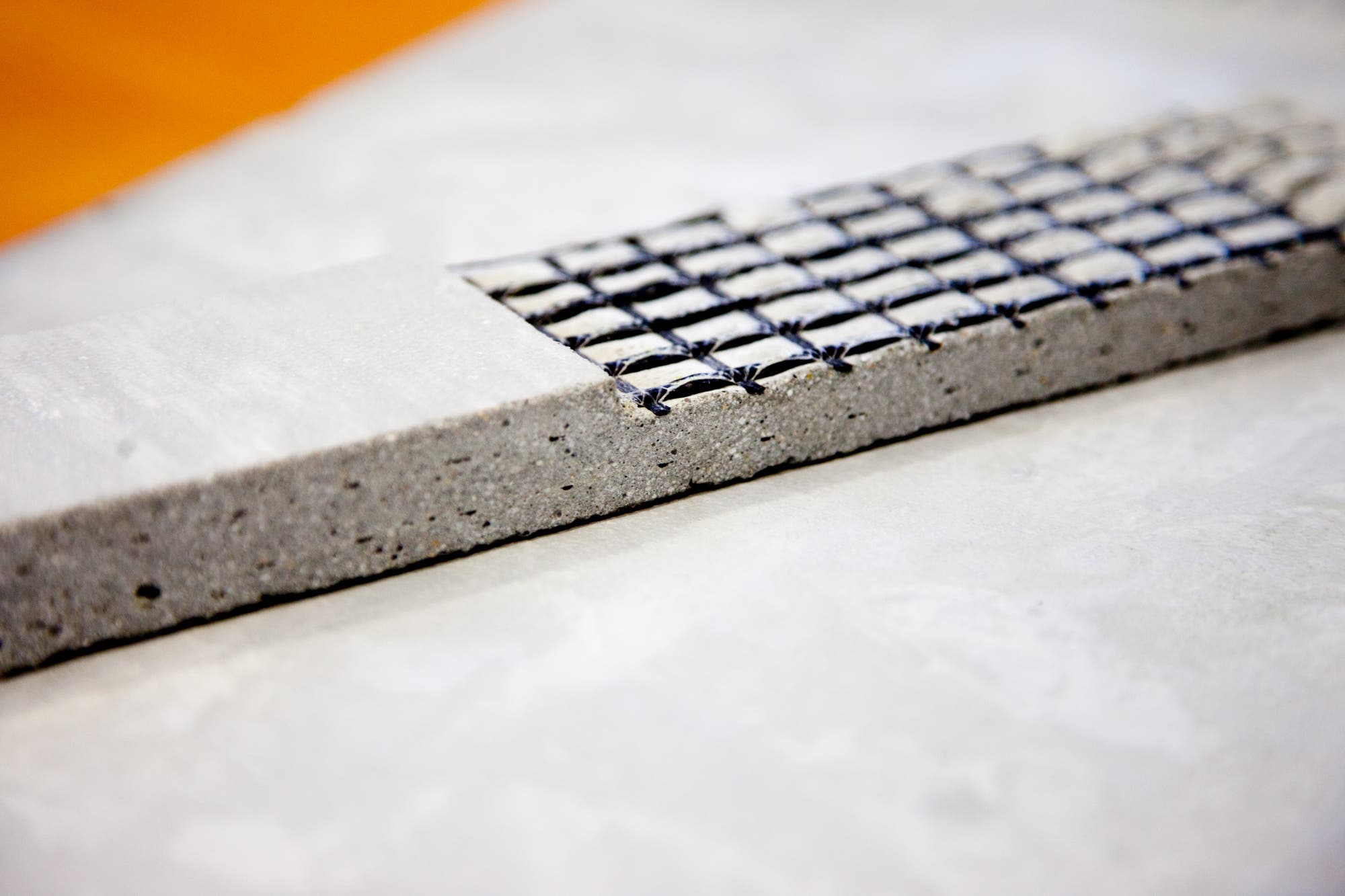 Ein rechteckiges Stück Beton mit einer eingebetteten Gitterstruktur aus schwarzem Material. Das Gitter ist teilweise sichtbar und verläuft parallel zur Oberfläche des Betons. Der Beton ist glatt und hat kleine Poren. Das Bild zeigt eine Nahaufnahme, die die Details der Materialien und deren Zusammensetzung hervorhebt.