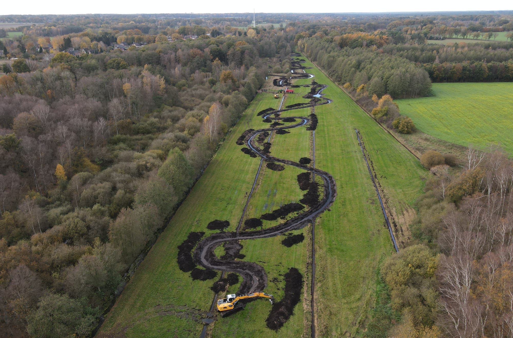 Luftaufnahme einer Landschaft mit einem neu angelegten, geschwungenen Wasserlauf, der sich durch ein grünes Feld zieht. Ein gelber Bagger ist am unteren Rand des Bildes zu sehen und arbeitet am Ufer des Wasserlaufs. Auf beiden Seiten des Feldes befinden sich dichte Wälder. Im Hintergrund sind vereinzelte Häuser und eine weite Landschaft zu erkennen. Die Szene vermittelt den Eindruck von Landschaftsgestaltung und Renaturierung.