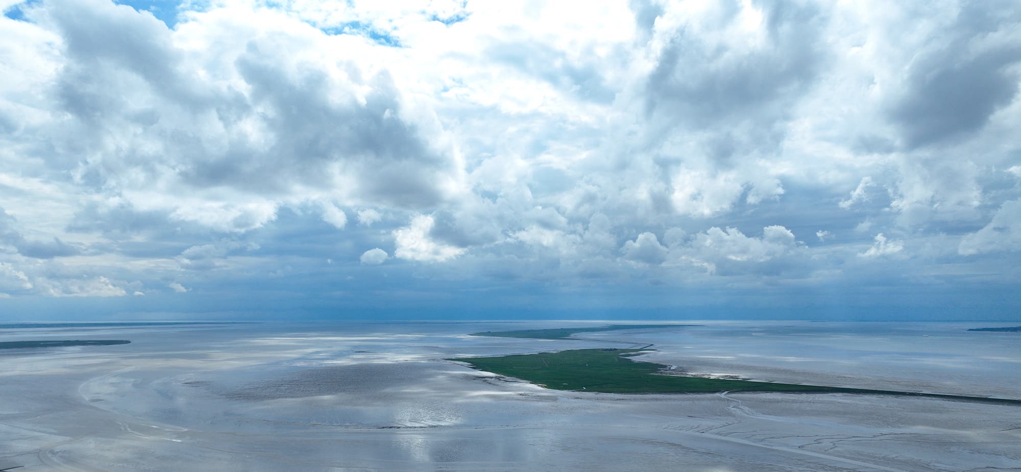 Weitläufige Küstenlandschaft mit einer grünen Landzunge, die sich in das ruhige, graue Wasser erstreckt. Der Himmel ist mit dramatischen, weißen Wolken bedeckt, die das Licht durchscheinen lassen. Keine Menschen oder Gebäude sind sichtbar.
