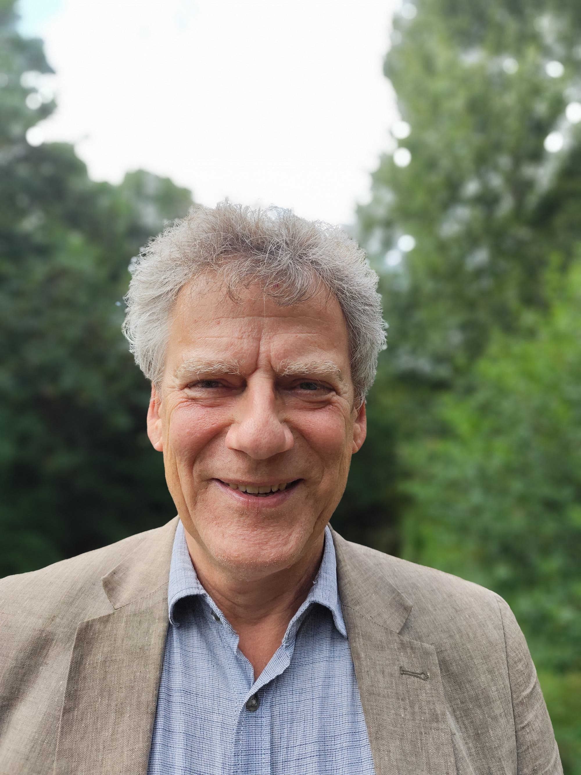 Ein lächelnder Mann in einem beigen Sakko und einem blauen Hemd steht im Freien vor einer grünen, verschwommenen Waldkulisse. Der Himmel ist hell und leicht bewölkt.