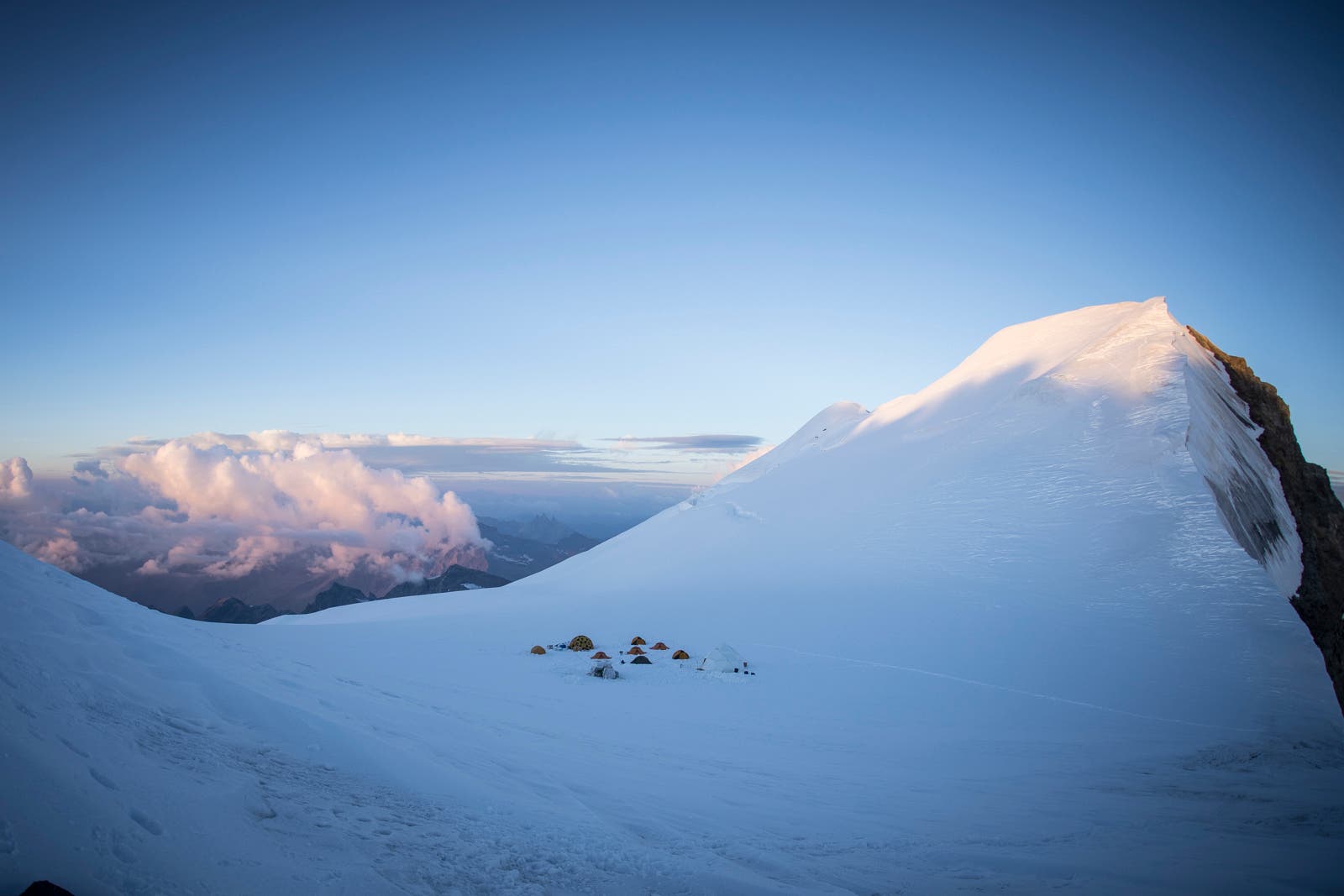 Blick af einen gletscherbedeckten Berghang, der wie eine Skischanze in die Höhe ragt. vorn auf dem Gletscher ein Camp von Zelten.