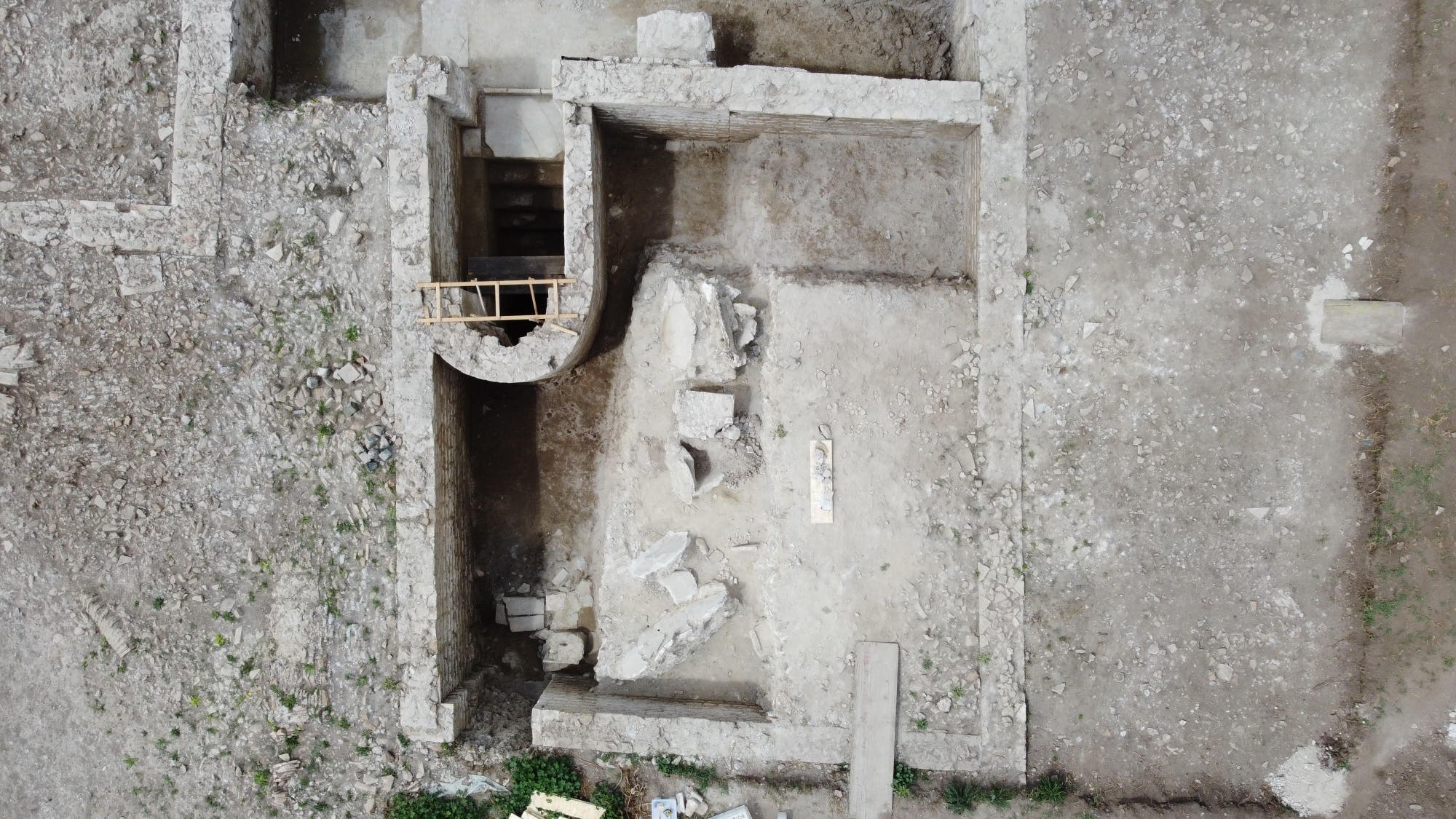 Luftaufnahme einer archäologischen Ausgrabungsstätte mit frei gelegten Mauerresten und einem rechteckigen Grundriss. In der Mitte befindet sich eine halbkreisförmige Struktur mit einer kleinen Holzleiter darauf. Es handelt sich um die Reste eines jüdischen Ritualbads. Der Boden ist uneben und von Erde und Steinen bedeckt. Keine Personen sind sichtbar.