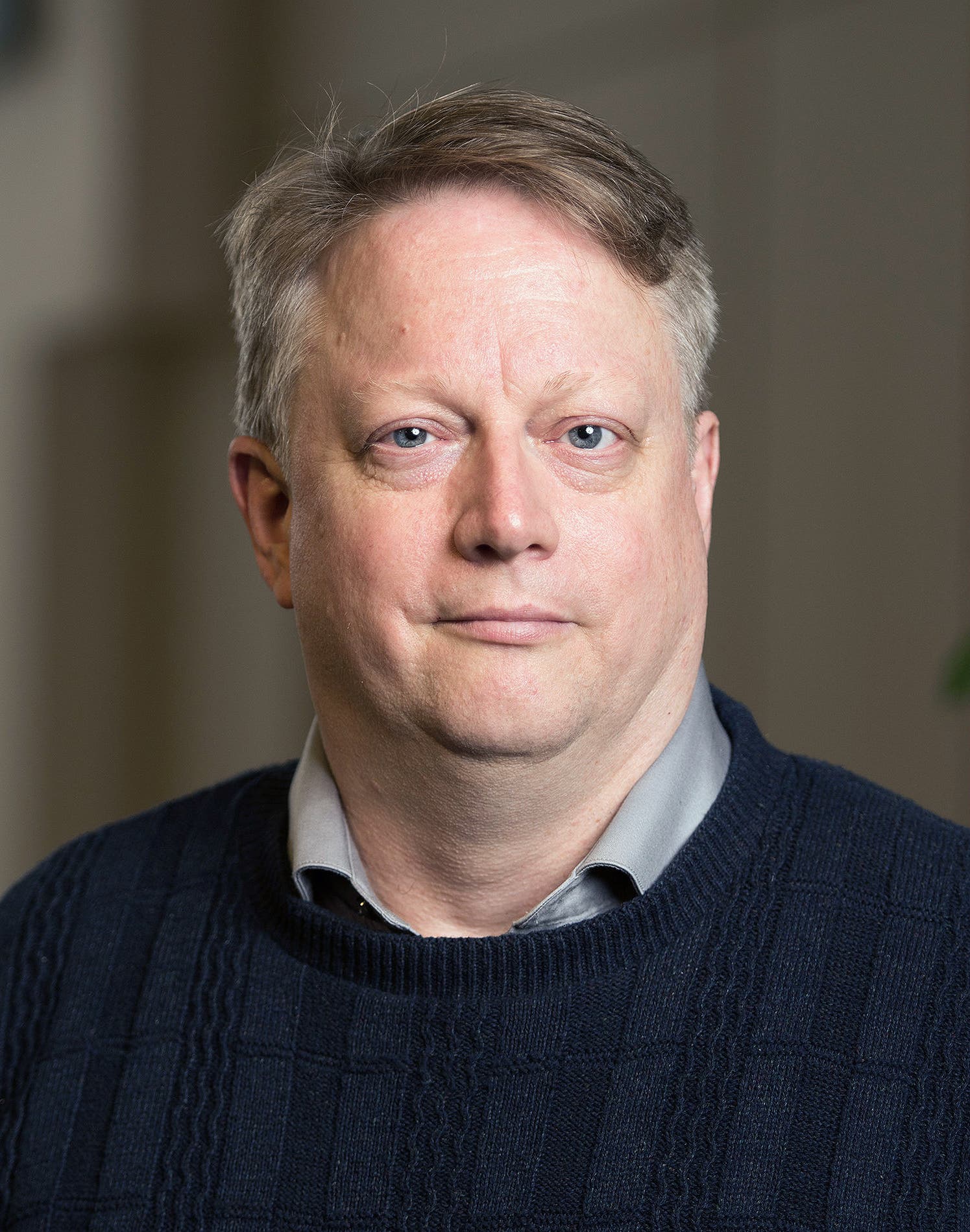 Der Sozialpsychologe David Dunning blickt direkt in die Kamera. Er hat kurzes, helles Haar und trägt einen dunkelblauen Pullover und ein graues Hemd. Der Hintergrund ist unscharf und neutral.