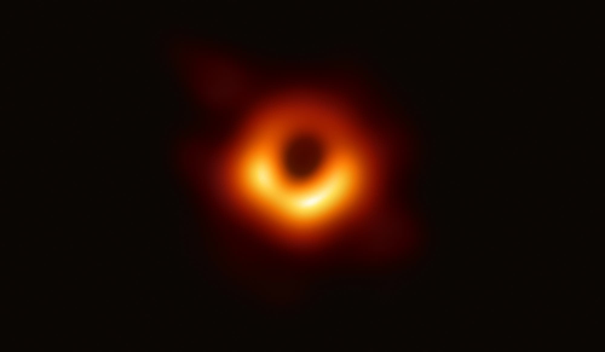 Ein verschwommenes, ringförmiges Bild eines Schwarzen Lochs, das von einem hellen, leuchtenden Ring aus Gas und Staub umgeben ist. Der Hintergrund ist dunkel, was den Kontrast zum leuchtenden Ring verstärkt. Dieses Bild zeigt das erste direkte visuelle Abbild eines Schwarzen Lochs, aufgenommen vom Event Horizon Telescope. Schlüsselwörter: Schwarzes Loch, Event Horizon Telescope, Astronomie, leuchtender Ring.