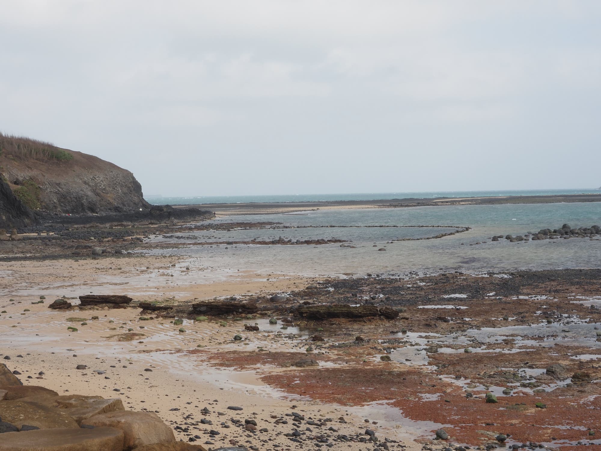 Küstenlandschaft bei Ebbe mit felsiger Klippe links und sandigem Strand im Vordergrund. Der Himmel ist bewölkt, und das Meer erstreckt sich bis zum Horizont. Die frei gelegten Felsen und Algen auf dem Sand deuten auf einen niedrigen Wasserstand hin. Die Szene vermittelt eine ruhige, natürliche Atmosphäre.