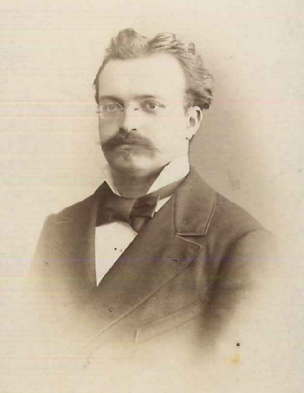 Ein historisches Schwarz-Weiß-Foto (Sepia) von Ernst Hartwig in formeller Kleidung mit Anzug und Fliege. Er trägt eine Brille und hat einen gepflegten Schnurrbart. Der Hintergrund ist neutral und schlicht, was den Fokus auf die Person lenkt.