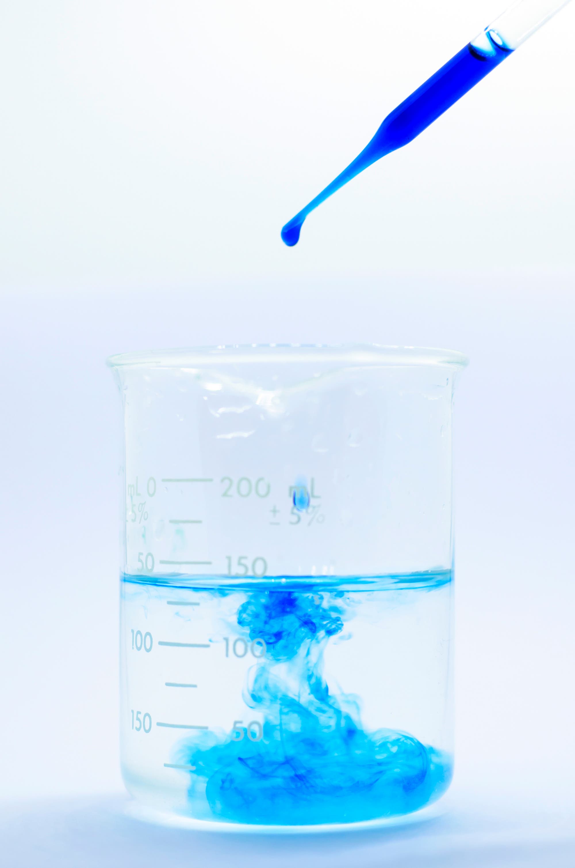 Aus einer Pipette tropft blaue Tinte in ein Becherglas mit klarem Wasser und verteilt sich