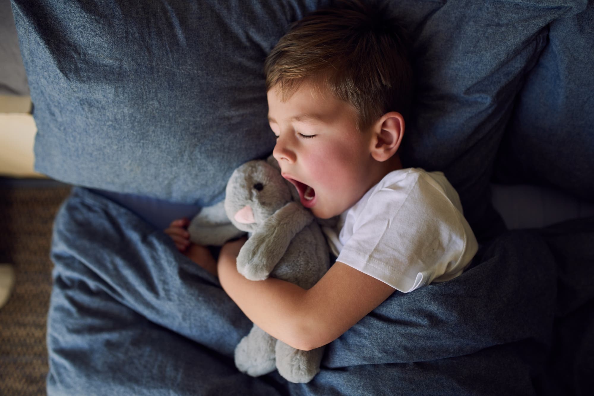 Ein Junge liegt mit seinem Kuscheltier im Bett und gähnt