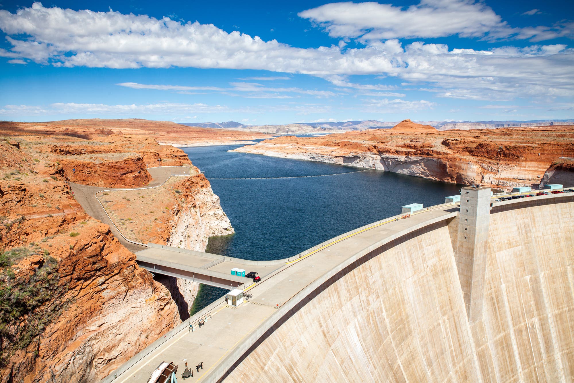 Eine große Betonstaumauer erstreckt sich über einen tiefen Canyon mit rotem Gestein, durch den ein breiter Fluss fließt. Der Himmel ist klar mit einigen Wolken. Auf der Staumauer sind mehrere Personen zu sehen, die die Aussicht genießen. Eine Straße führt über die Mauer und verbindet die beiden Seiten des Canyons. Im Hintergrund erstreckt sich die Landschaft mit weiteren Felsformationen.